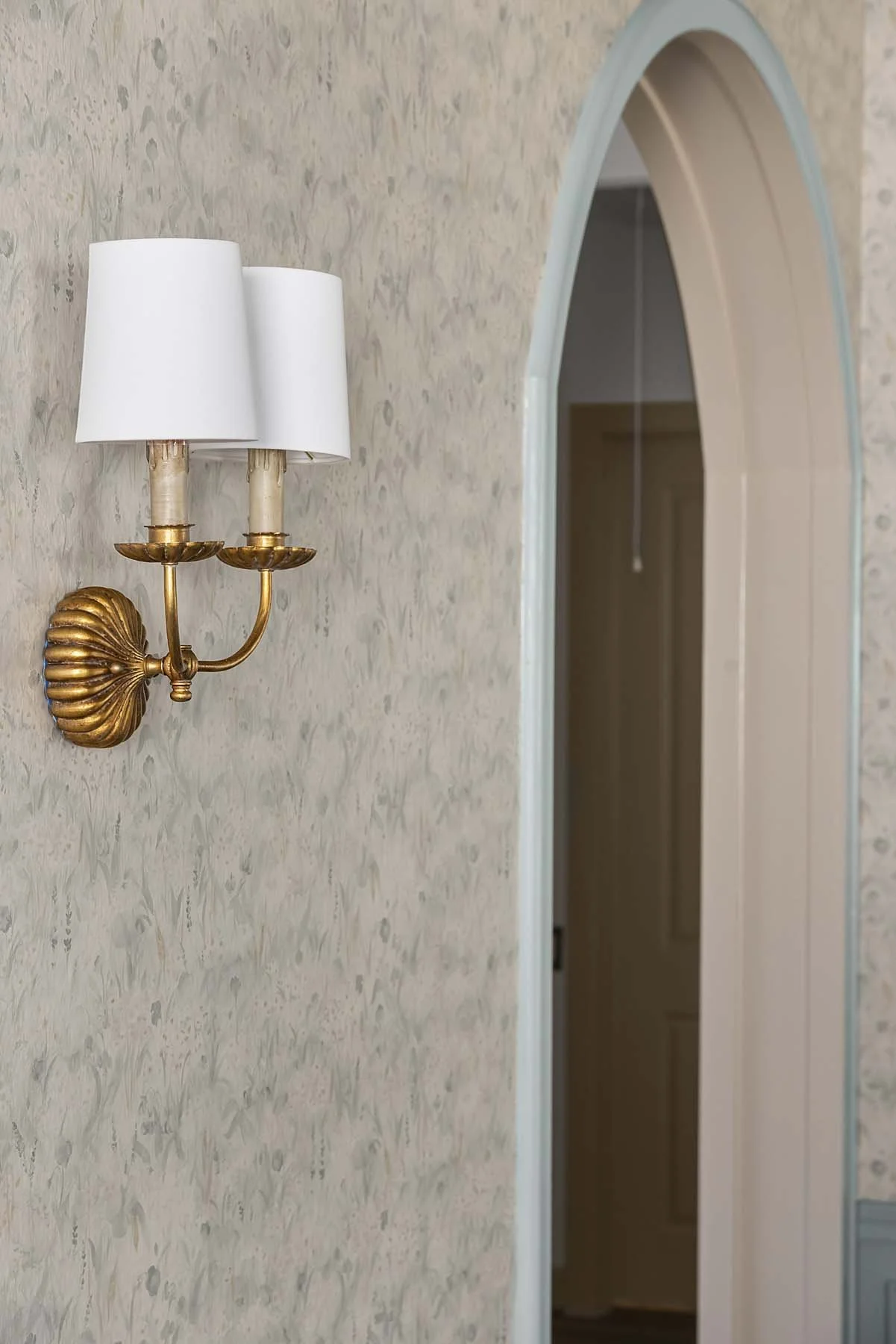 A gold-colored double wall sconce with white lampshades mounted on a textured, pastel-colored wall, next to an arched doorway.
