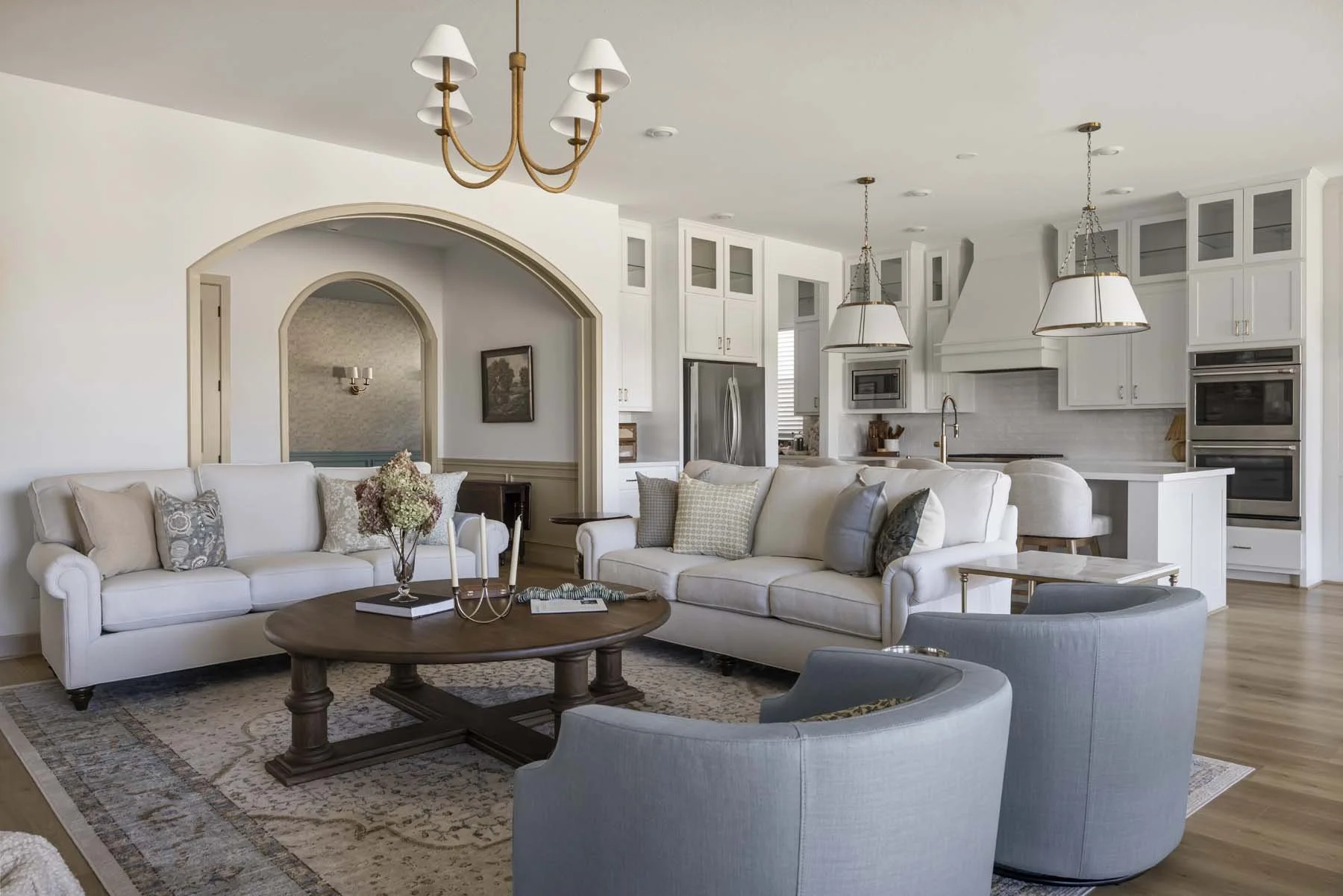 Open-concept living room and kitchen with white sofas, wooden coffee table, gray armchairs, chandelier, and modern kitchen with white cabinetry and stainless steel appliances.
