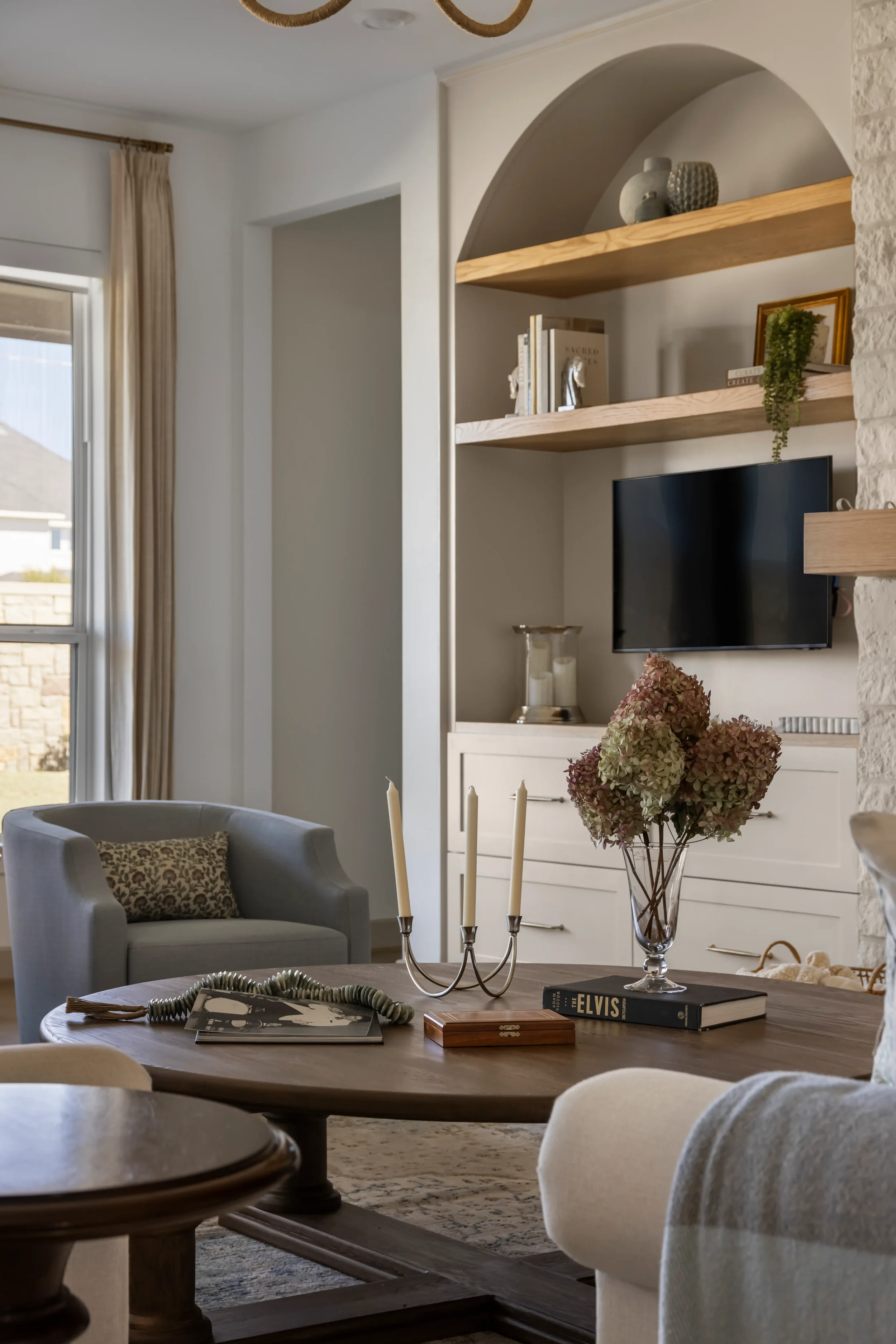 Living room with a wooden coffee table, floral centerpiece, books, and a candle holder. A TV mounted on a white wall with built-in shelves holding books and decorative items. Light-colored armchairs and a window with beige curtains.