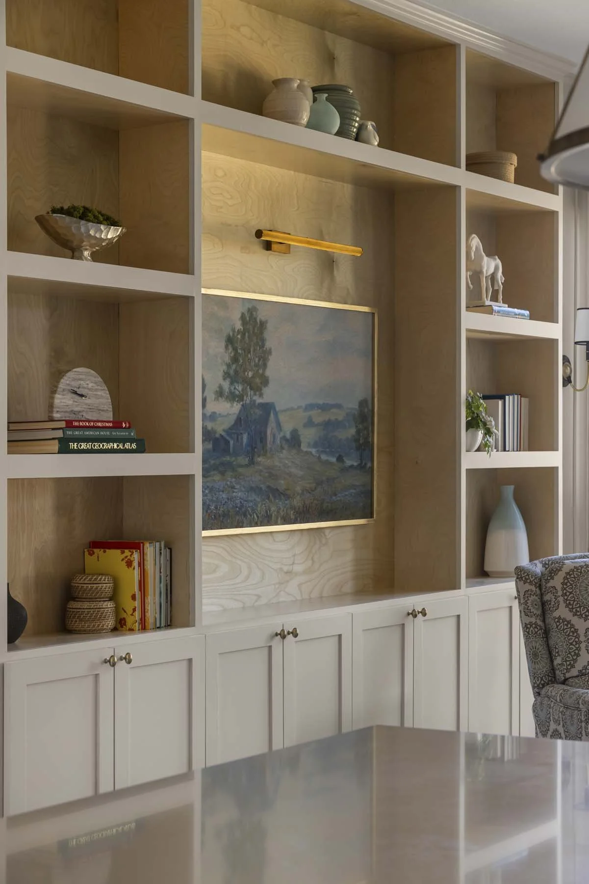 Wood-paneled wall with built-in white shelving, displaying books, vases, and decorative objects, with a painting of a rural landscape and a reading light