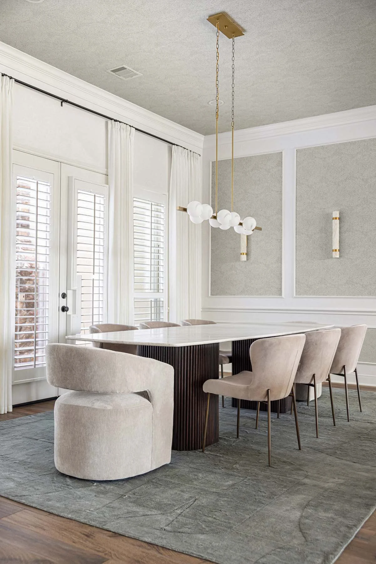 A modern dining room with a large white marble table, surrounded by beige upholstered chairs on a gray area rug, with large windows covered by white curtains and shutters, and a unique chandelier hanging overhead.