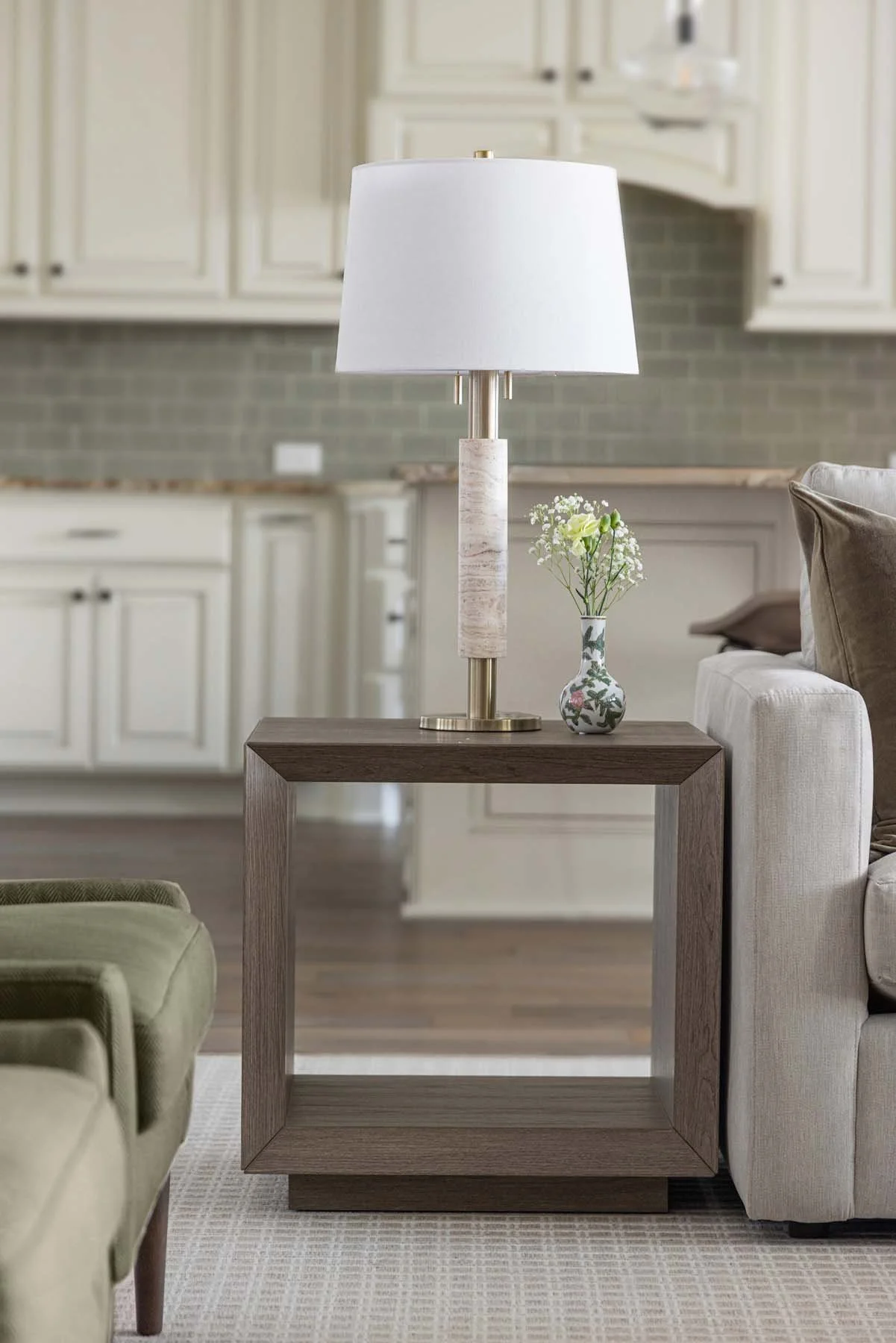 A living room with a wooden side table holding a white table lamp and a small vase with white flowers, surrounded by beige and green seating.