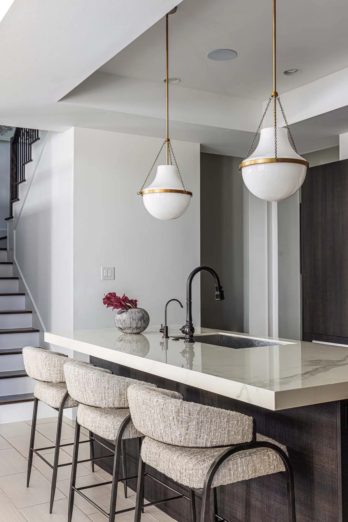Modern kitchen with a cream-colored marble island, black faucet, three beige upholstered barstools, two hanging pendant lights, and a dark cabinet in the background. A vase with dark red flowers is on the island.
