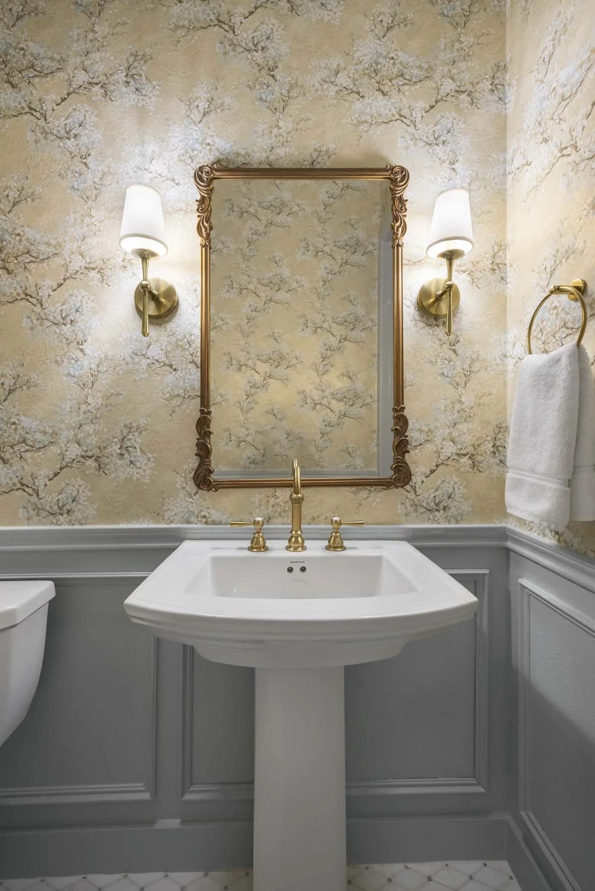A bathroom with a pedestal sink, gold fixtures, a large mirror with a decorative gold frame, and two wall-mounted light fixtures with white shades. There is a towel ring with a white towel on the right side.