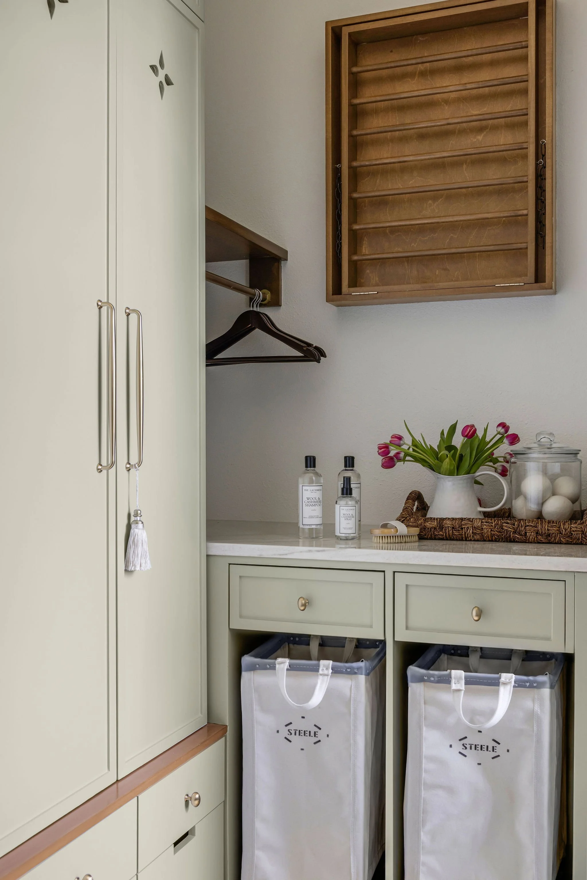 A laundry room corner with a cream-colored cabinet, a wooden shoe rack, and two fabric laundry hampers labeled 'Steele'. On the white countertop, there are bottles of laundry supplies, a vase with pink tulips, and a glass jar filled with white balls.