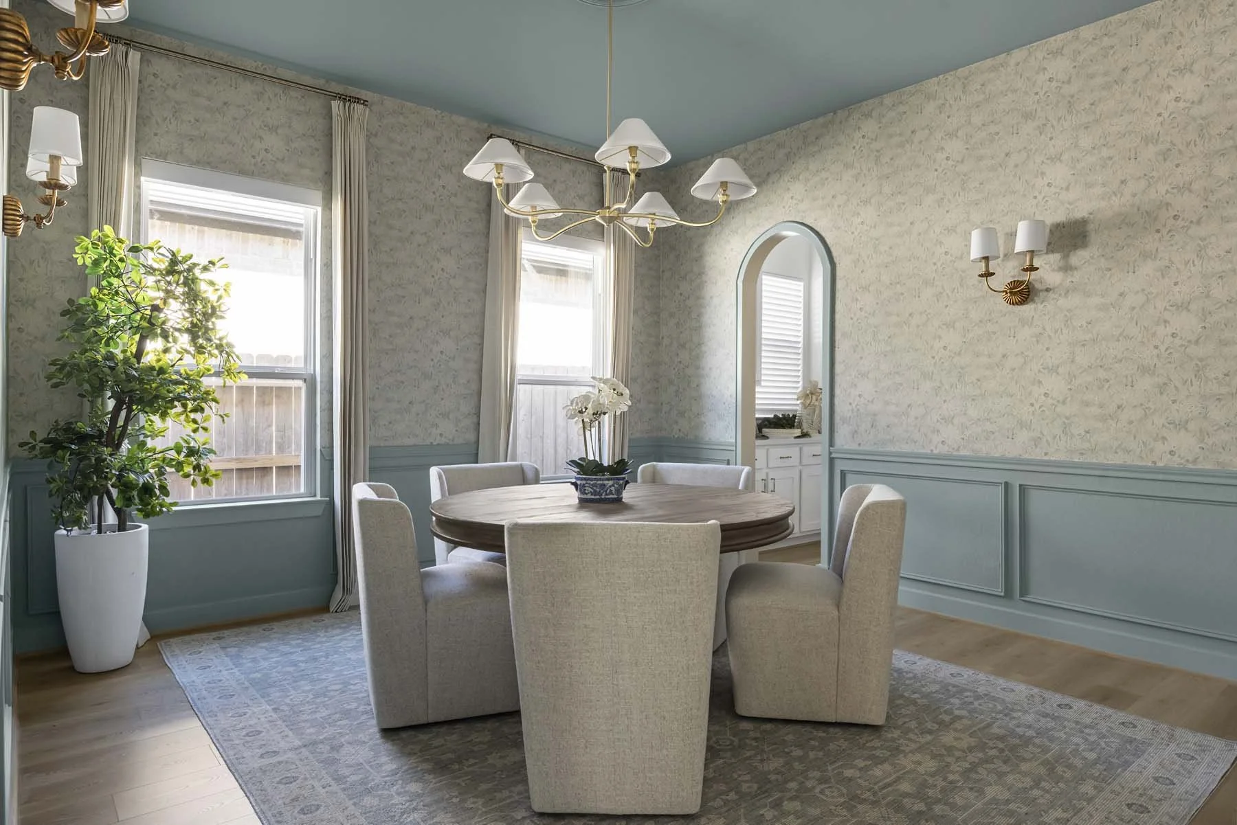 A dining room with a round wooden table surrounded by six upholstered chairs. There are two large windows with cream curtains, a potted plant to the left, and a chandelier hanging from the ceiling. A vase with white orchids is on the table, and the r