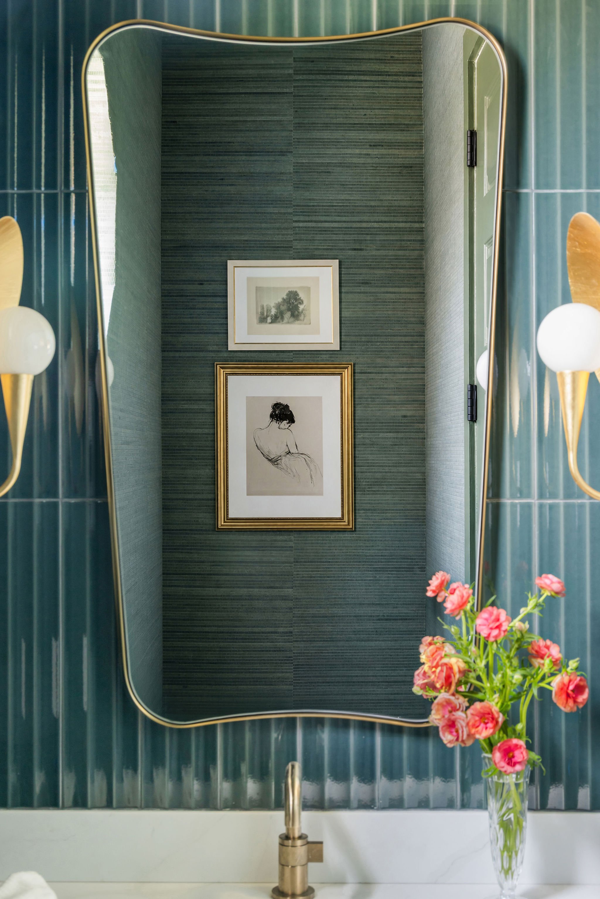 A bathroom vanity with a round mirror, a modern faucet, and a bouquet of pink flowers in a clear glass vase, with framed pictures on a dark textured wall in the background.