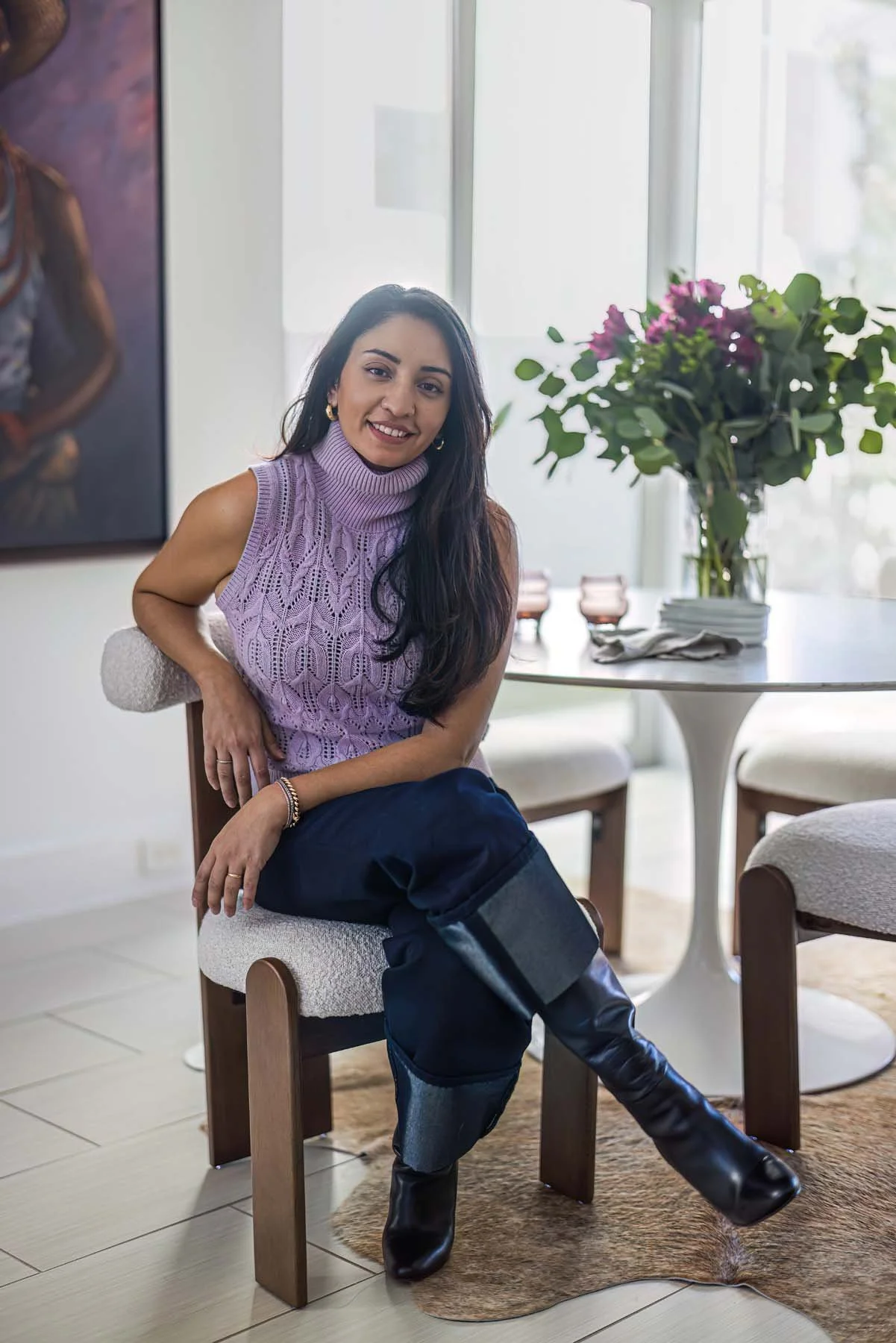 Woman sitting on a chair indoors, smiling, wearing a purple sleeveless knit top, dark jeans, and black knee-high boots, with a large bouquet of pink and purple flowers on a table nearby.