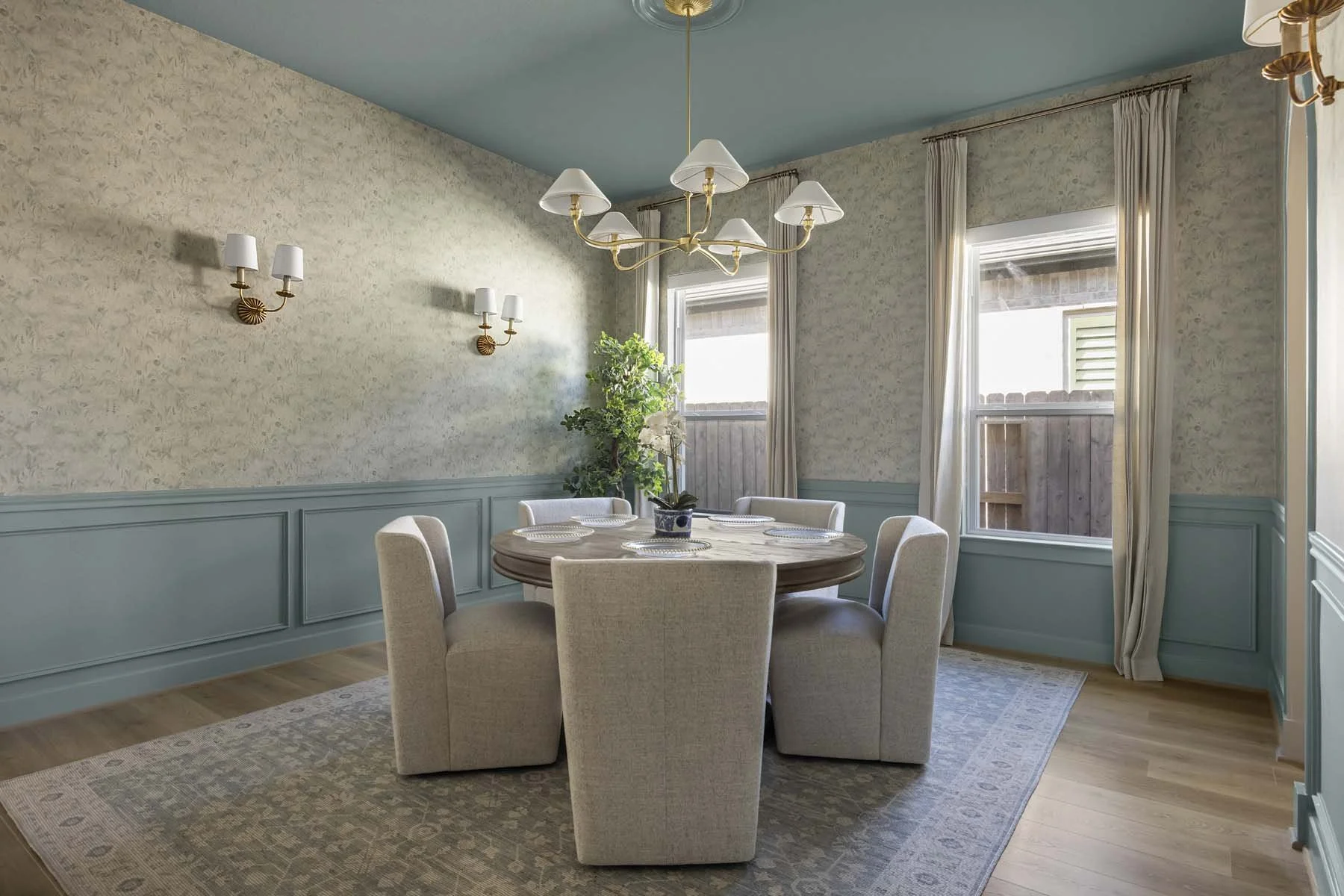 A dining room with a round wooden table set with four plates, surrounded by six upholstered beige chairs. The room has a light blue ceiling, cream floral wallpaper, wainscoting, and a chandelier with multiple lamps. Two windows with cream curtains le