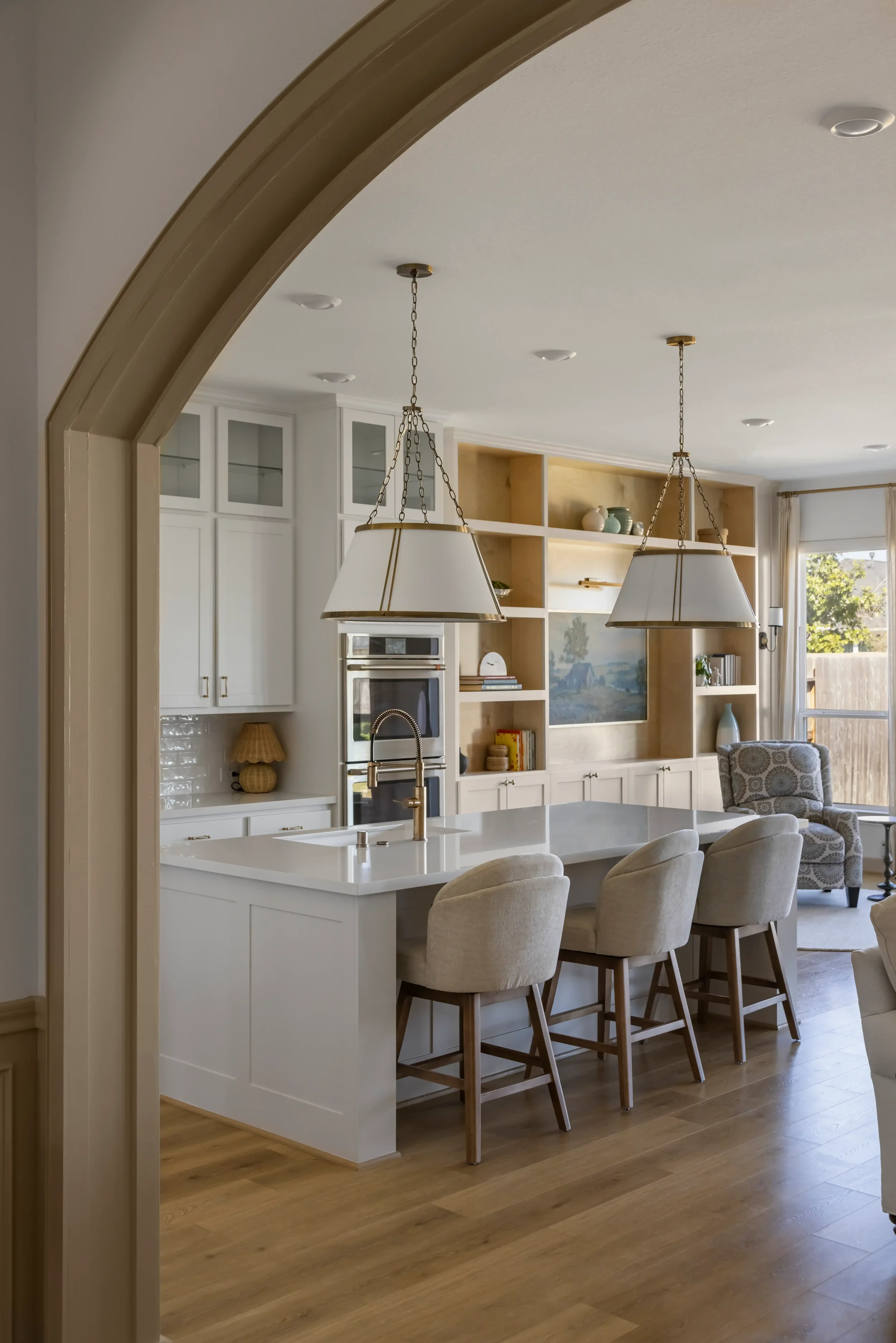 View of a bright, modern kitchen with white cabinets, a white island with a sink, and beige bar stools. Pendant lights hang above the island. The background features built-in shelves with decorative items and books, a double oven, and a window letting in natural light.