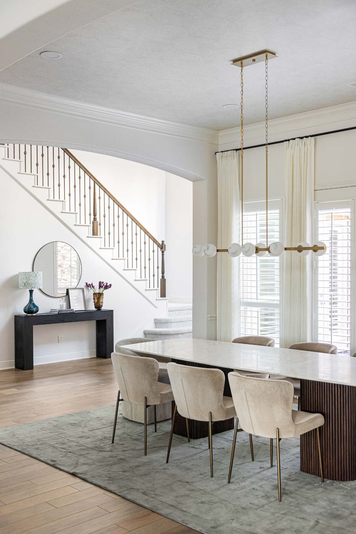Modern dining room with a marble-top table, six beige chairs, a geometric chandelier, and a staircase with a wooden banister in the background.