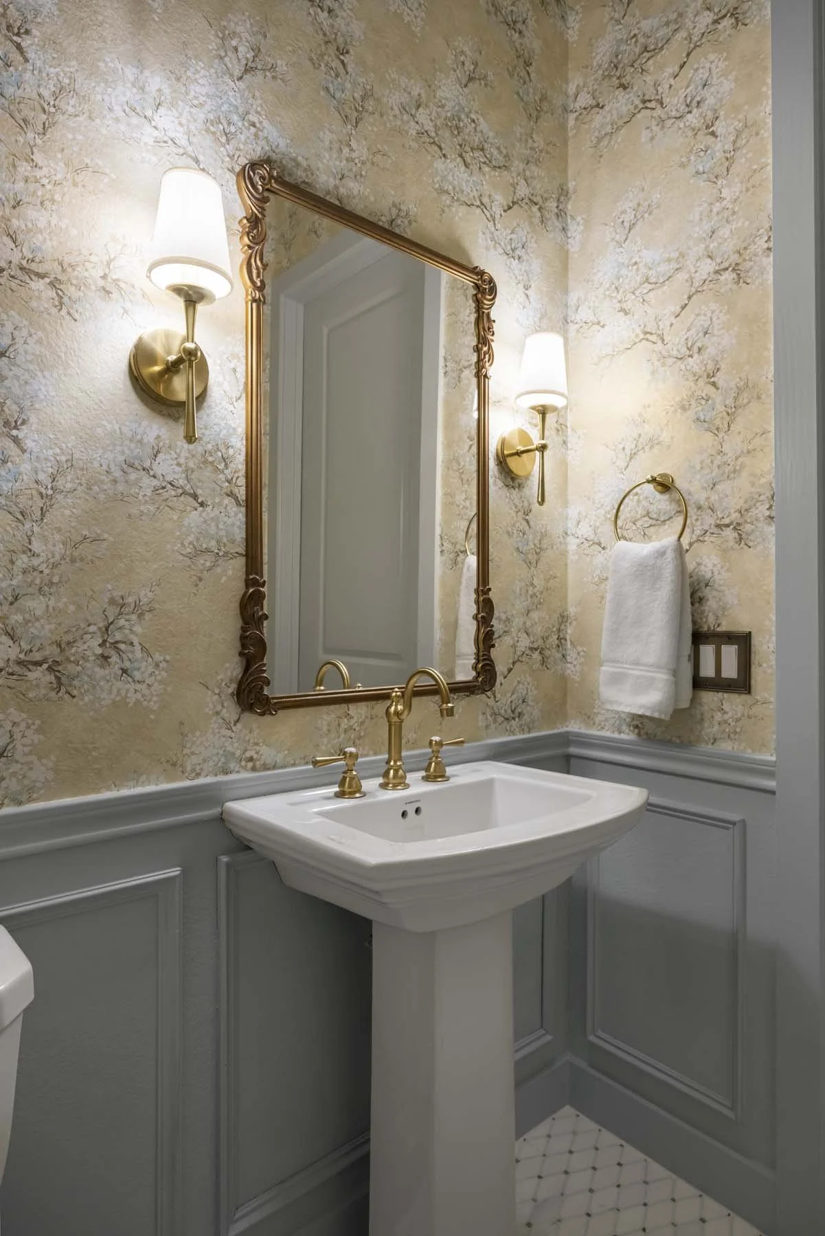 A traditional bathroom featuring a white pedestal sink with gold fixtures, a large ornate mirror, and two wall-mounted sconces with white shades. The walls are decorated with floral-patterned wallpaper, and a towel hangs on a gold ring next to a ligh