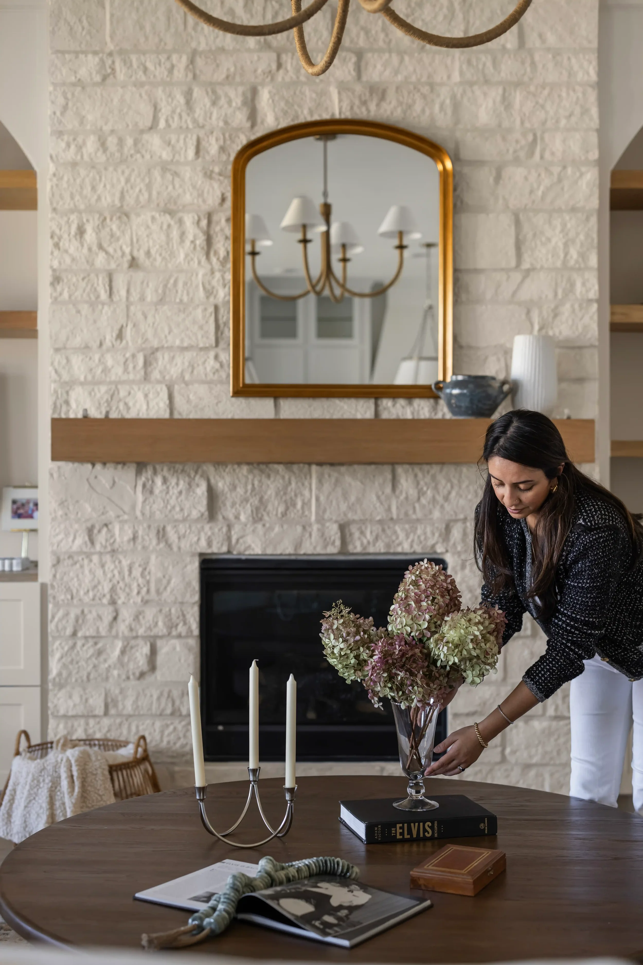 A woman arranging a bouquet of pink and green hydrangeas in a glass vase on a wooden dining table in front of a fireplace with a brick surround and a wooden mantel. On the table, there is a mailbox, an open magazine, a wooden box, and a silver candelabra with three white candles. A large mirror with a gold frame hangs above the fireplace, reflecting a chandelier with five lampshades.