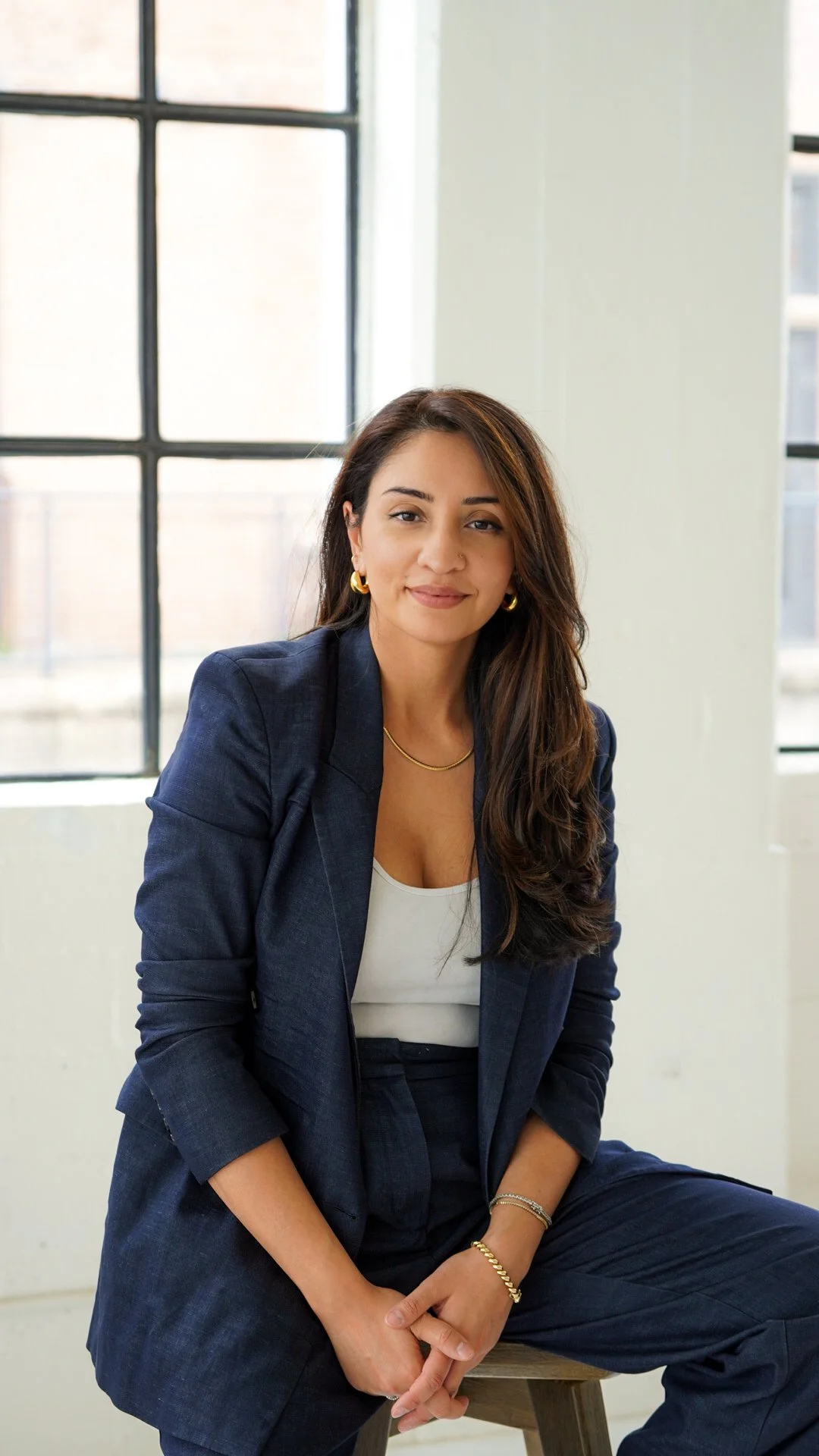 A woman in a dark blazer and pants sitting on a stool, with a large window in the background.
