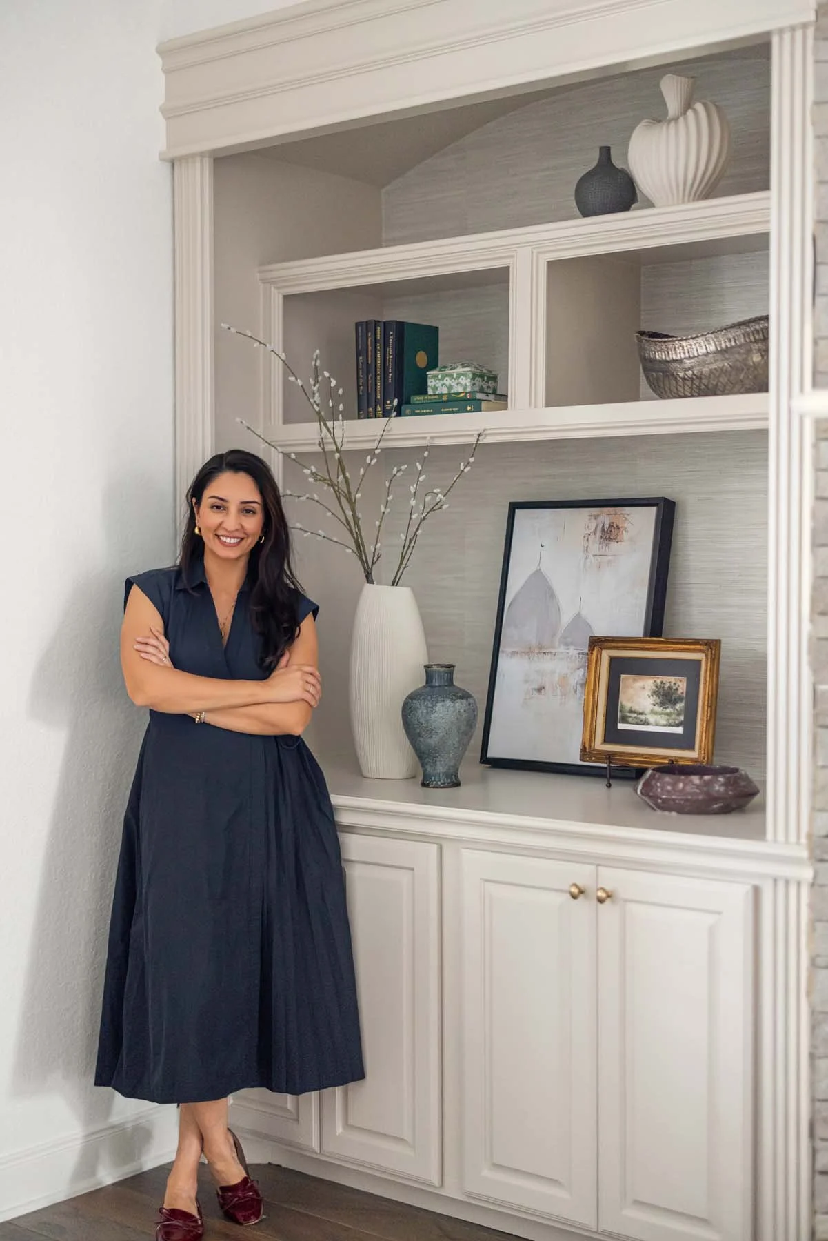 A woman with dark hair, wearing a navy dress and burgundy shoes, smiling and standing with arms crossed next to a white built-in shelf with decorative items and framed artwork.