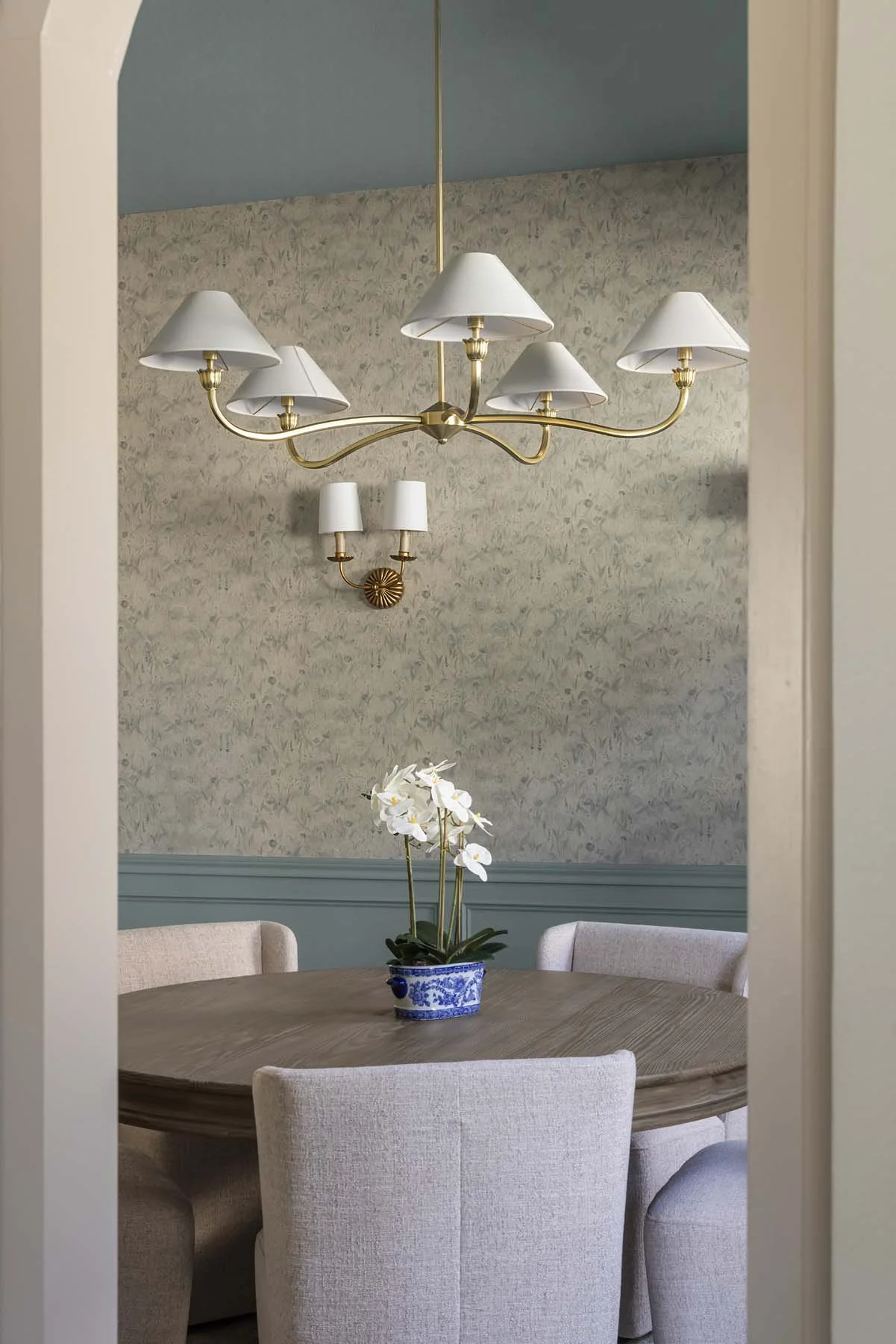 Elegant dining room with a wooden round table, four upholstered chairs, a white orchid in a blue and white ceramic pot, a gold chandelier with white lamp shades, and a wallpapered wall with blue wainscoting.