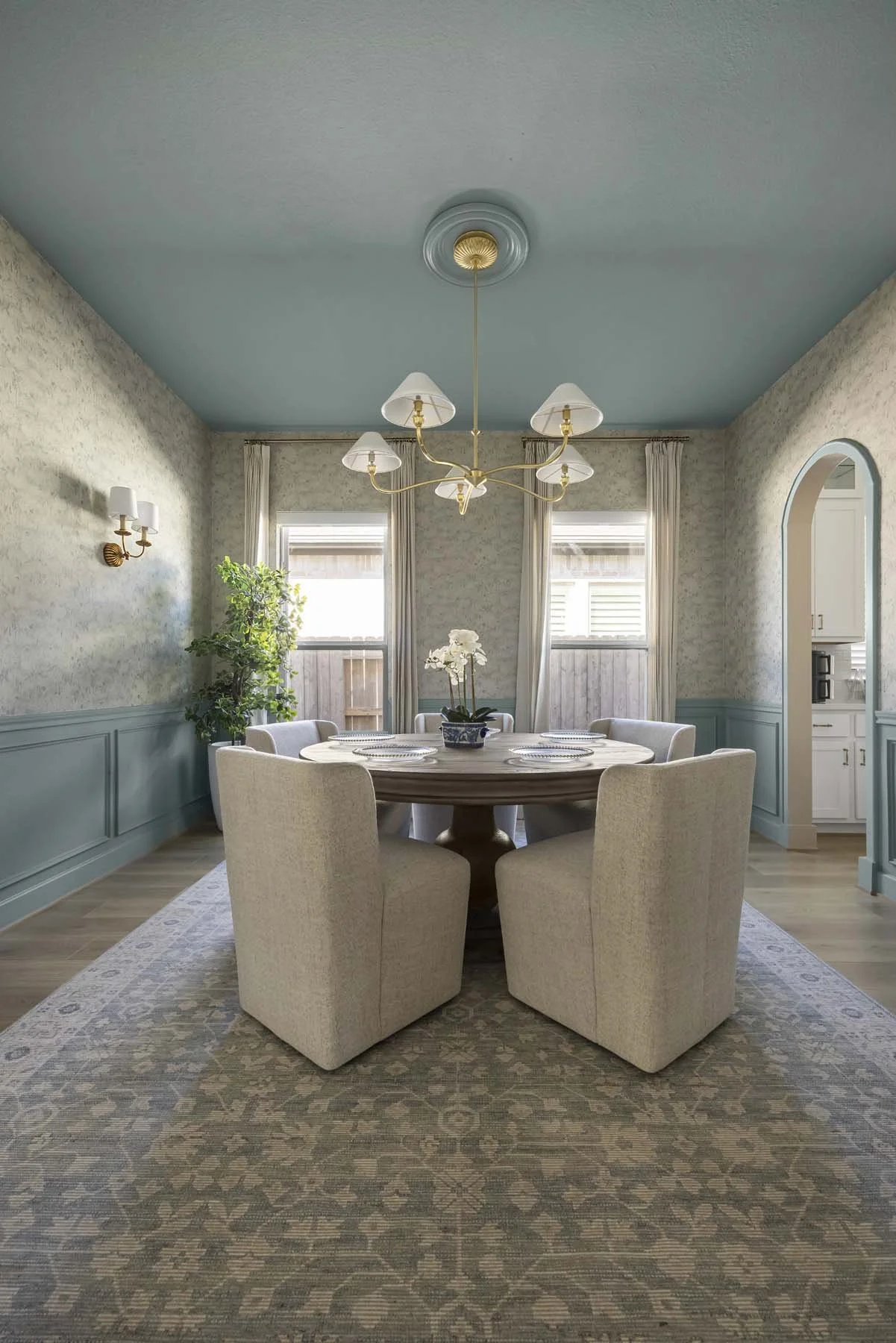 A dining room with a round wooden table surrounded by six beige upholstered chairs, a chandelier hanging from a blue ceiling, and two windows with white curtains.
