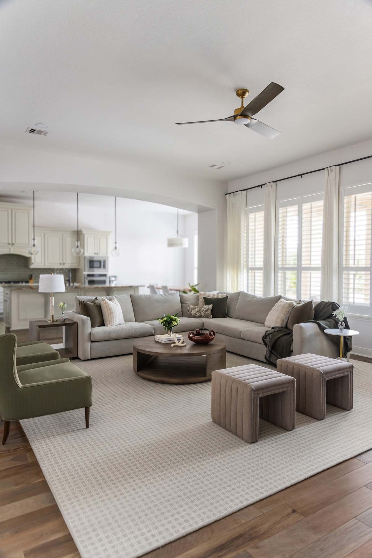 Living room with a large L-shaped beige sofa, green armchairs, wooden ottomans, and a round coffee table. There are large windows with white curtains letting in natural light, and a ceiling fan above.