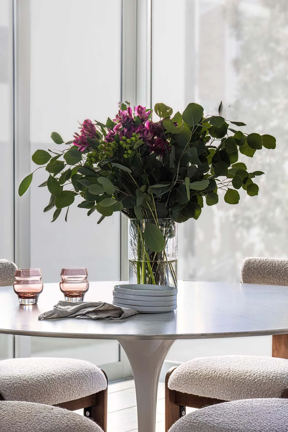 A round white marble table with a large glass vase holding a bouquet of pink and purple flowers and green foliage, two pink glasses, a stack of white plates, and a cloth, set in a bright room with large windows.