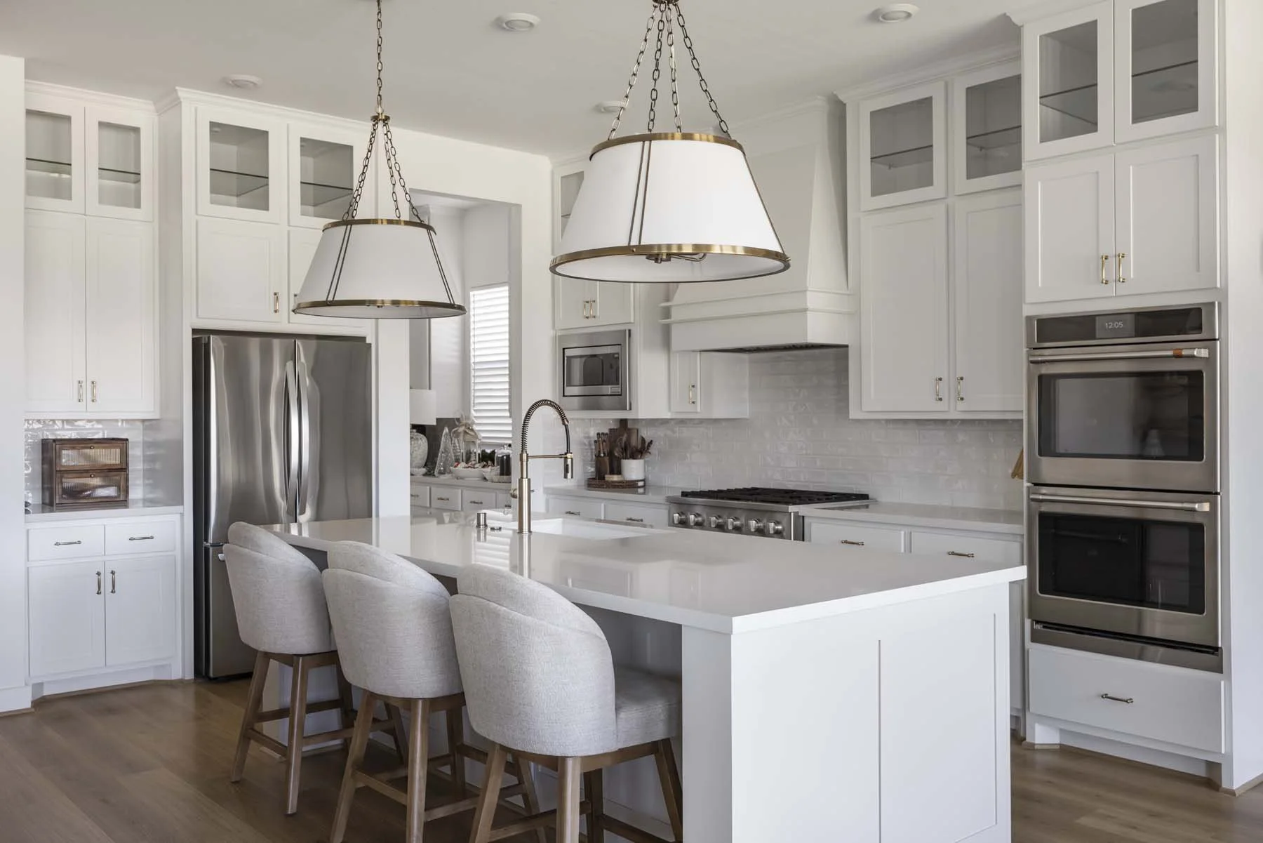 Modern white kitchen with a large island, three upholstered bar stools, stainless steel appliances, pendant lighting, cabinetry with glass doors, and hardwood flooring.
