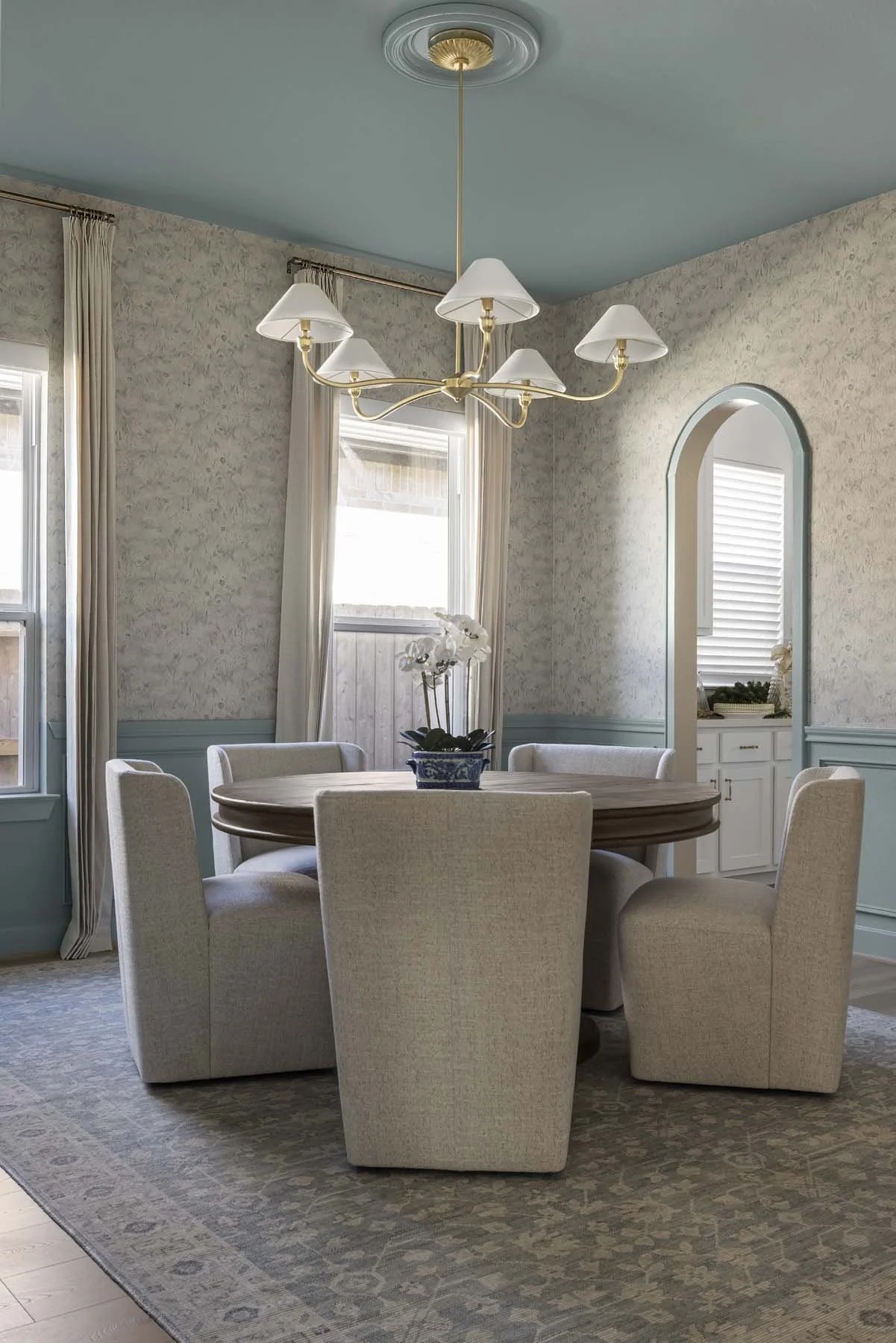 A dining room with a round wooden table, six upholstered chairs, chandelier, and a floral centerpiece. Two windows with curtains, a patterned rug, and a doorway with an arched frame.