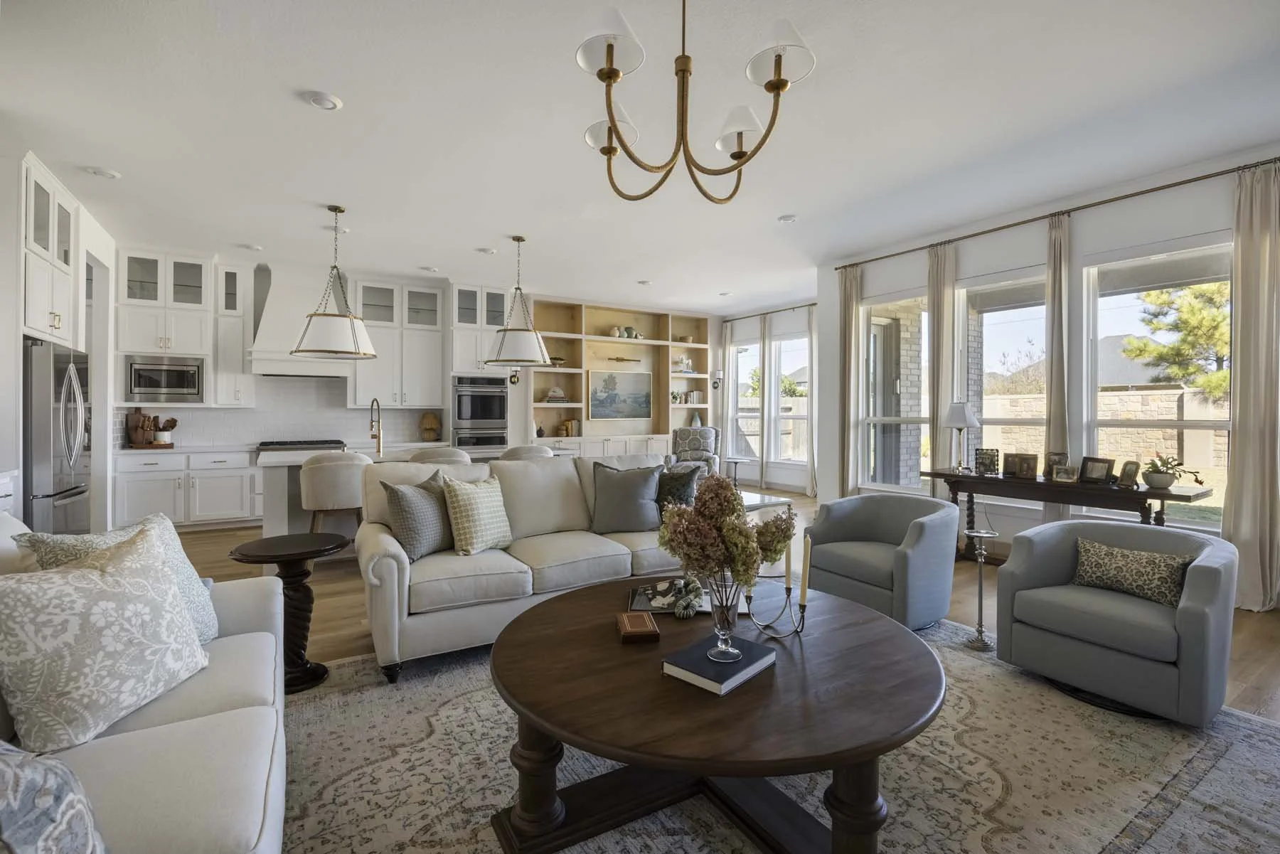 Bright open-concept living room and kitchen with white cabinets, large windows, beige sofas, and a wooden coffee table, decorated in a neutral color palette.