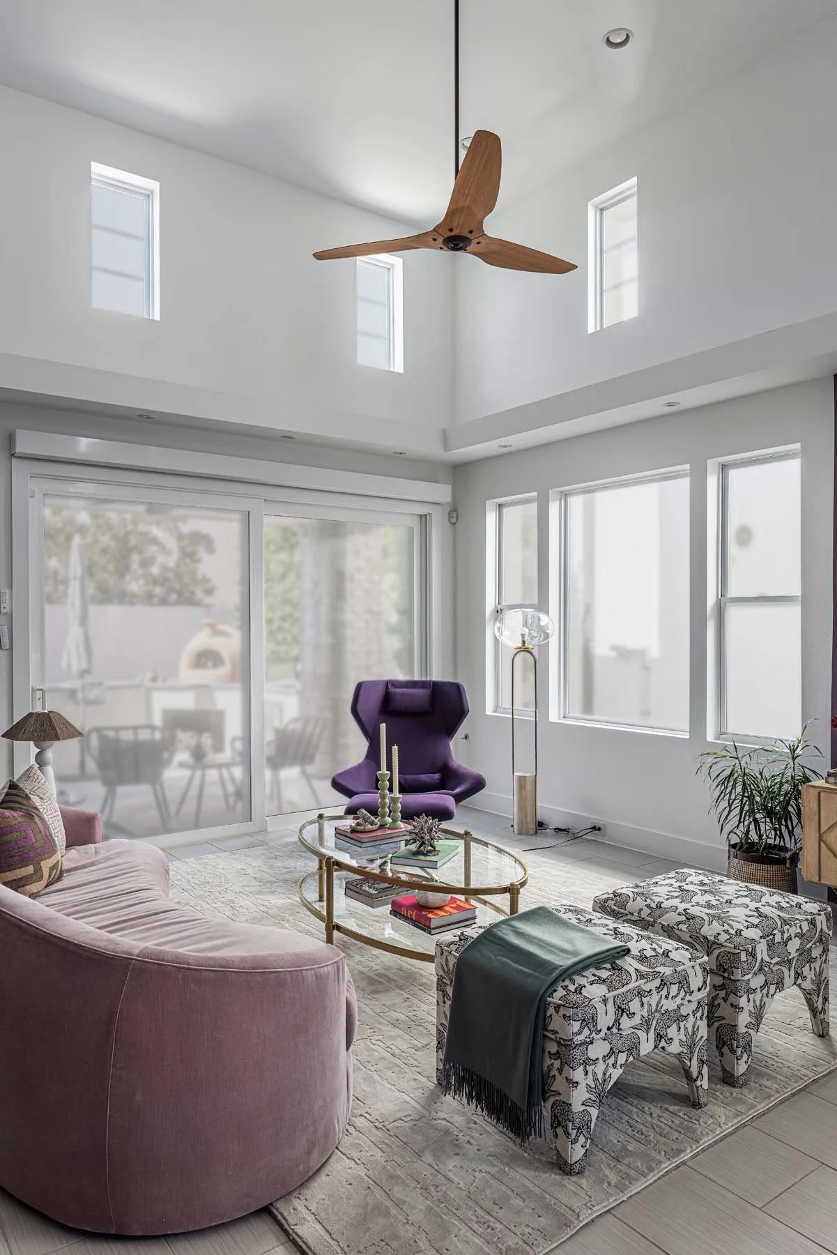 Living room with pink curved sofa, purple armchair, glass coffee table, patterned ottomans, and large windows with sliding door.