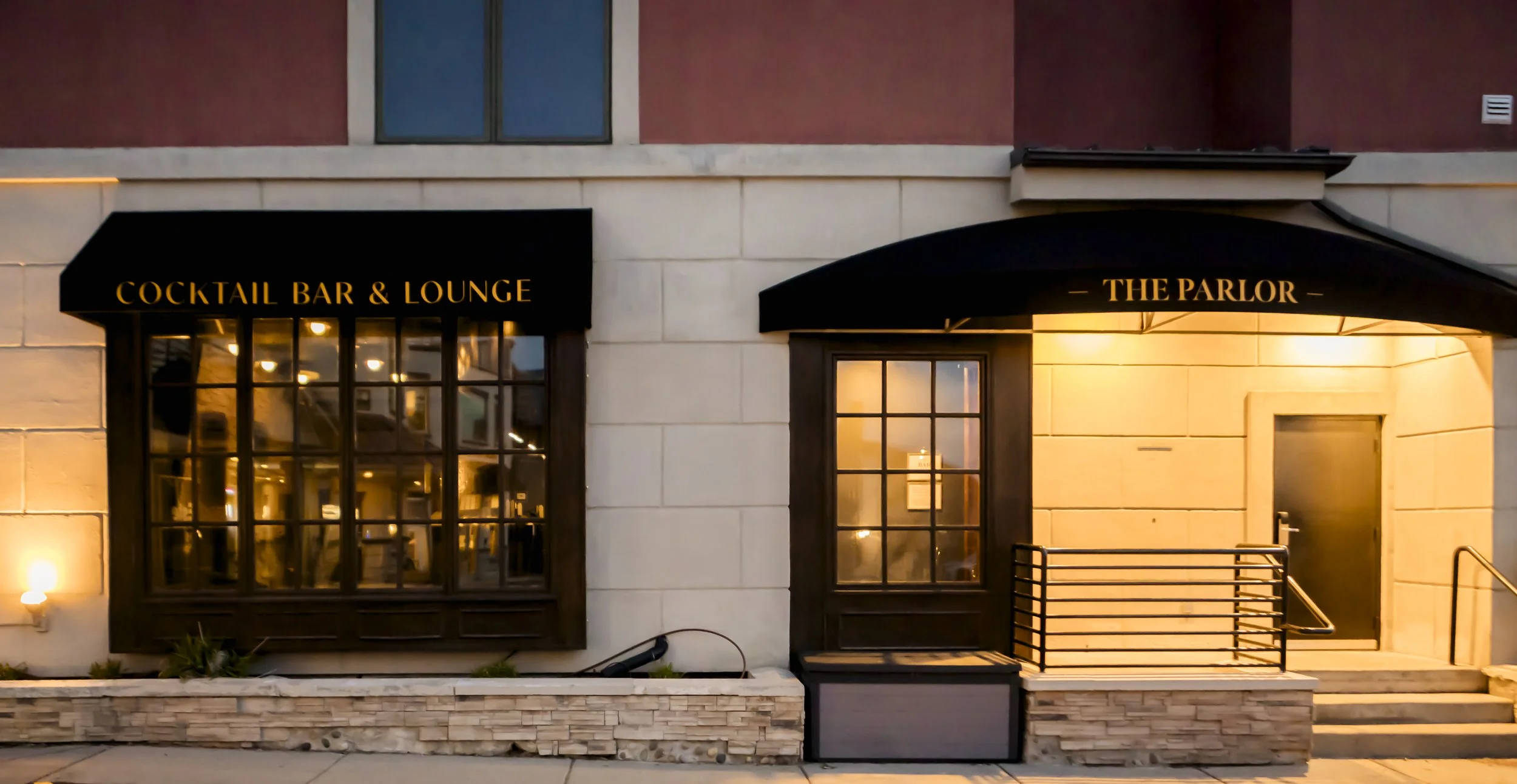 Exterior of a building with two black awnings labeled 'Cocktail Bar & Lounge' and 'The Parlor' in gold lettering, large windows, and a small staircase with a black railing.