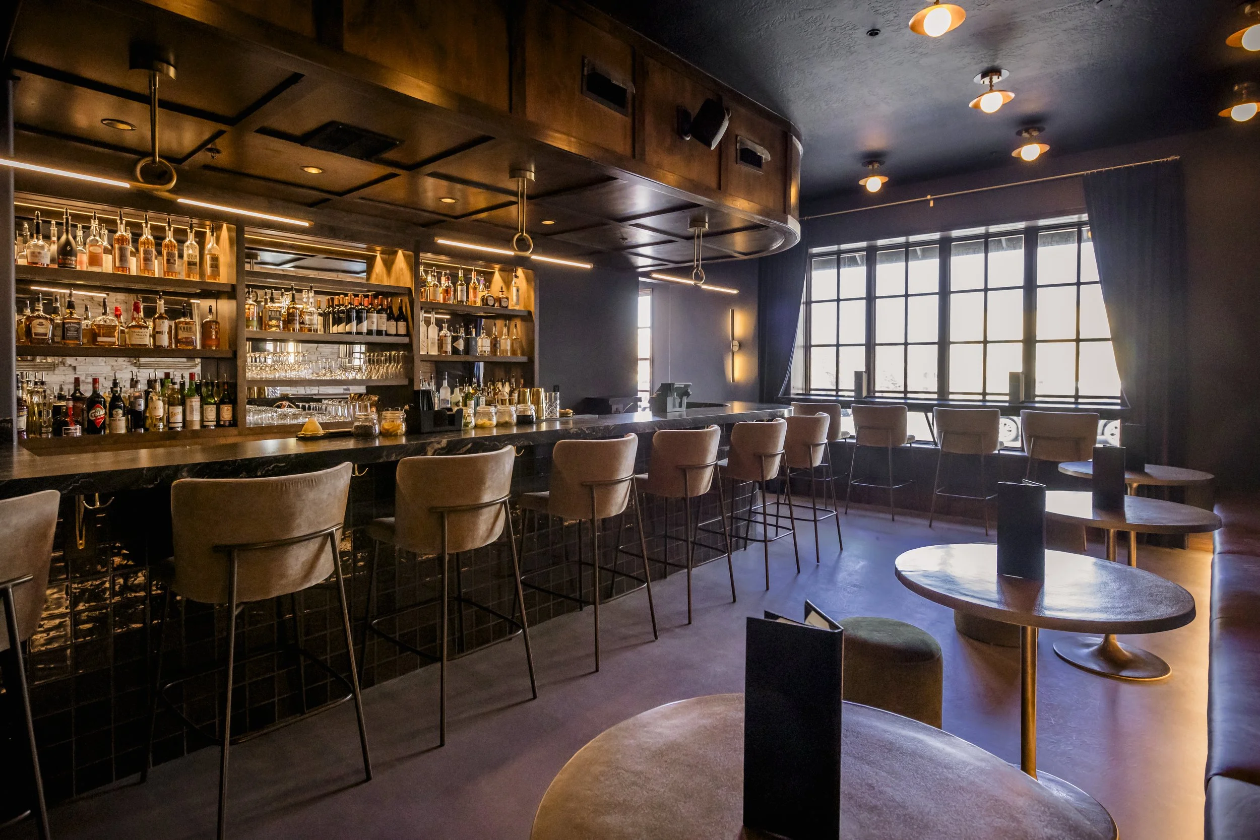 Interior of a modern bar with a long counter, shelves of various bottles of alcohol, bar stools, and large windows with dark curtains, illuminated by ceiling lights and warm lighting.