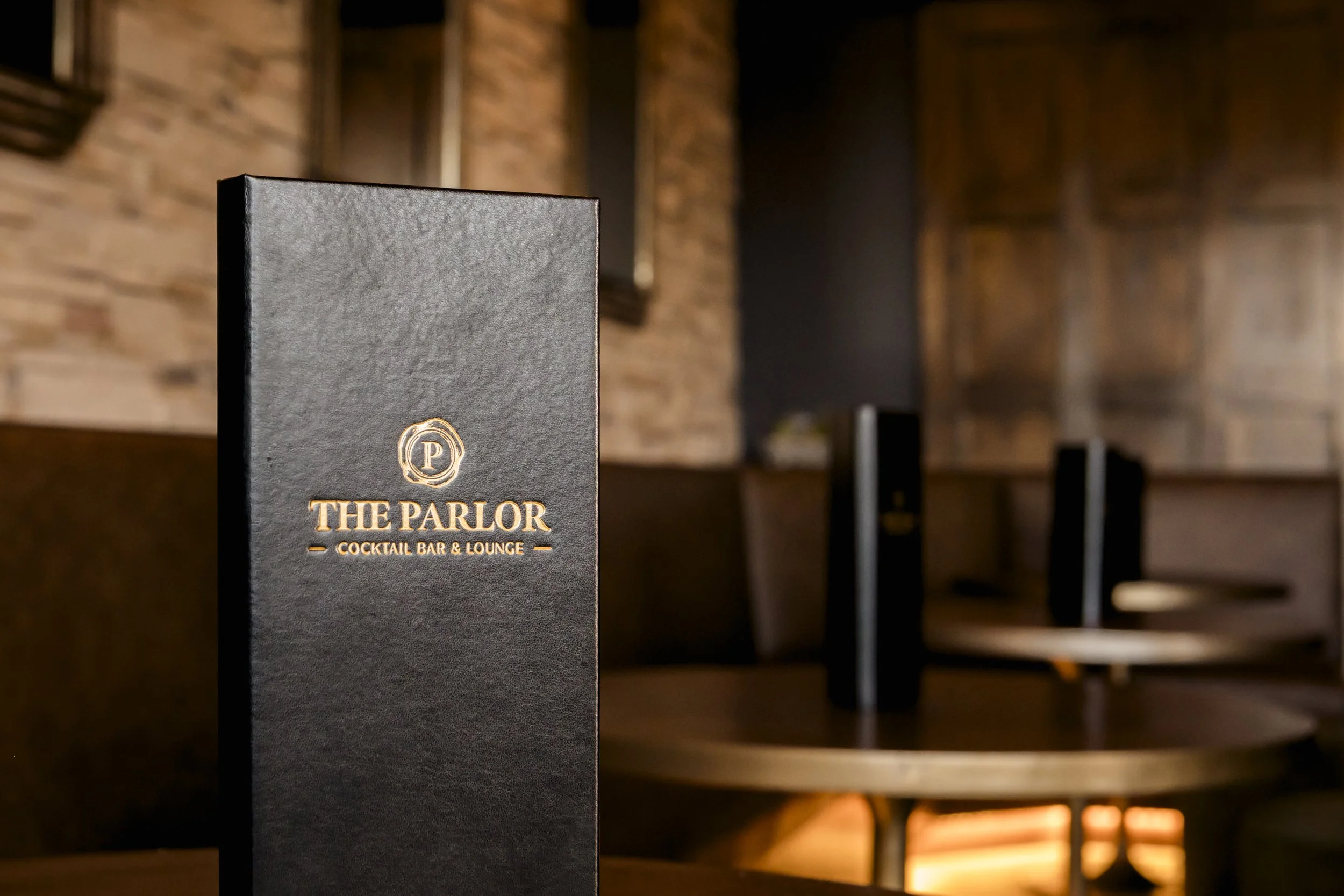 Close-up of a menu or flyer for The Parlor cocktail bar and lounge, with a blurred background of a dimly lit brick interior.