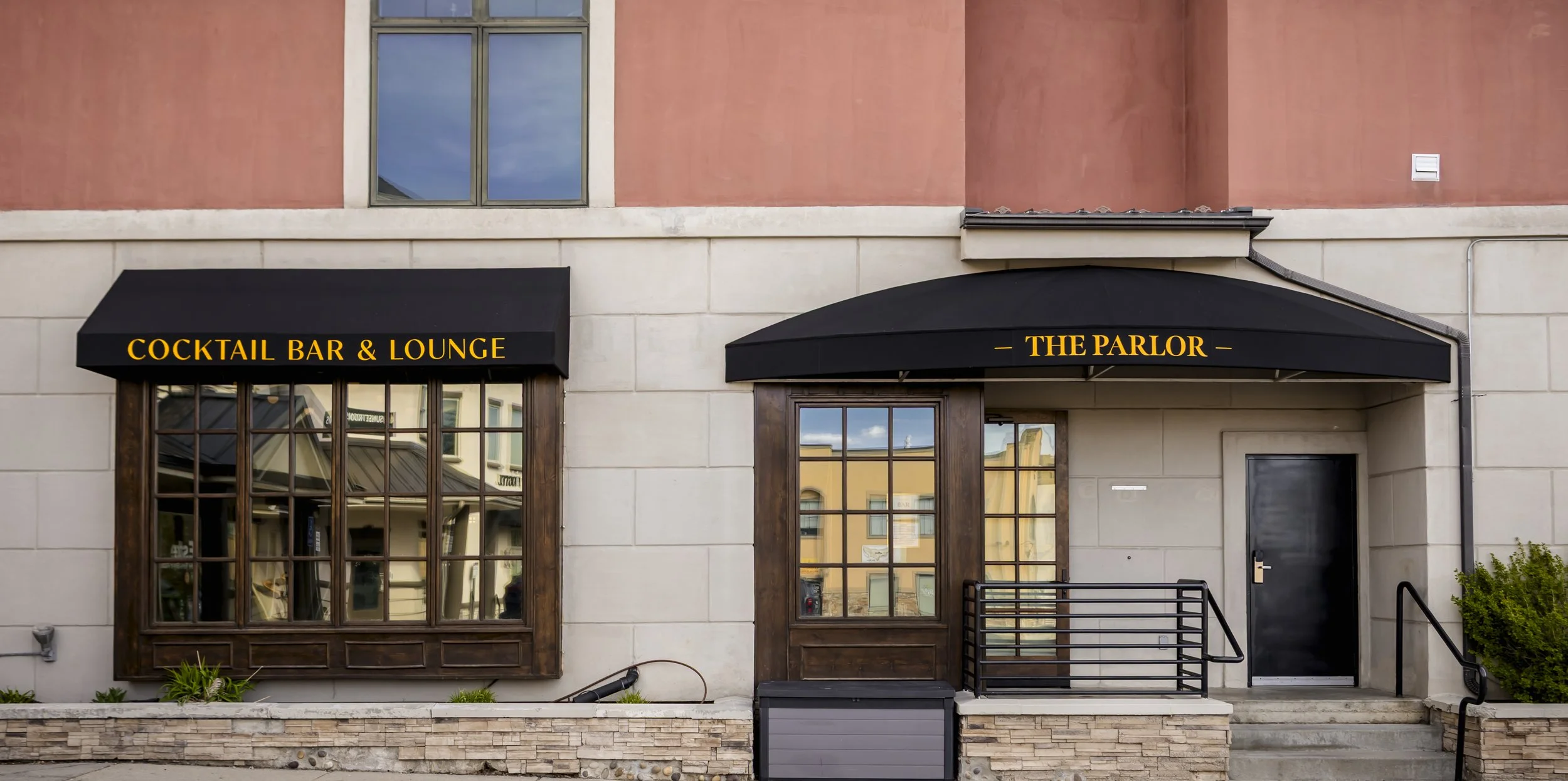 Exterior of a building with two black awnings; the left one reads 'Cocktail Bar & Lounge' and the right one reads 'The Parlor' in yellow letters. There are windows and a door beneath the awnings, with some reflection of the street scene visible in th