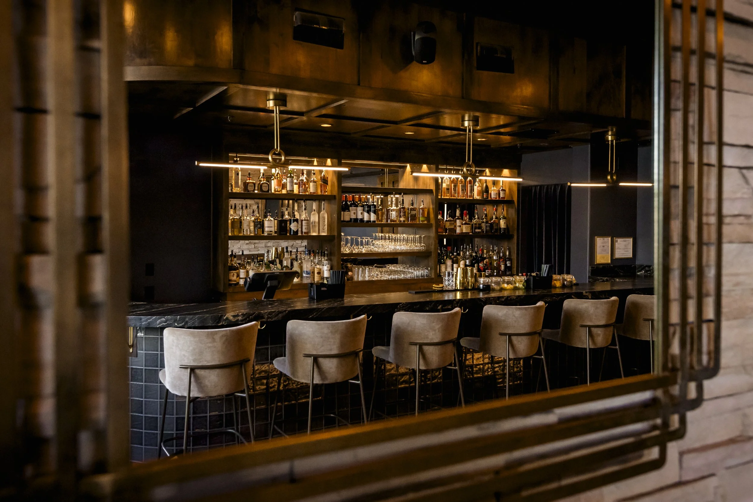 Interior view of a bar with a dark wooden shelf stocked with various bottles of alcohol and glasses, illuminated by warm lighting, with seating in front.