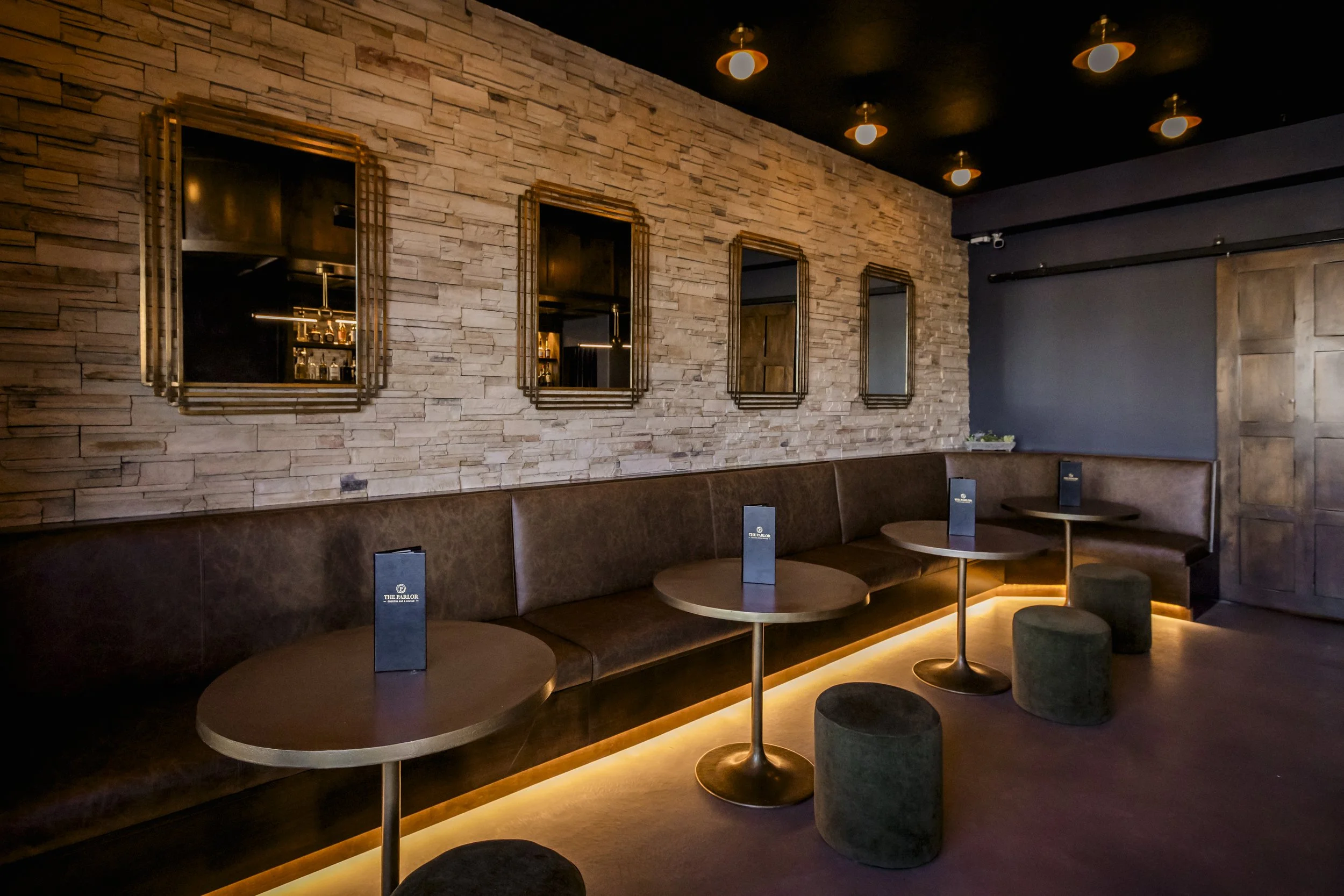 Interior of a lounge with a stone accent wall, round tables, cushioned seating, and mirrors with gold frames, illuminated by warm lighting.
