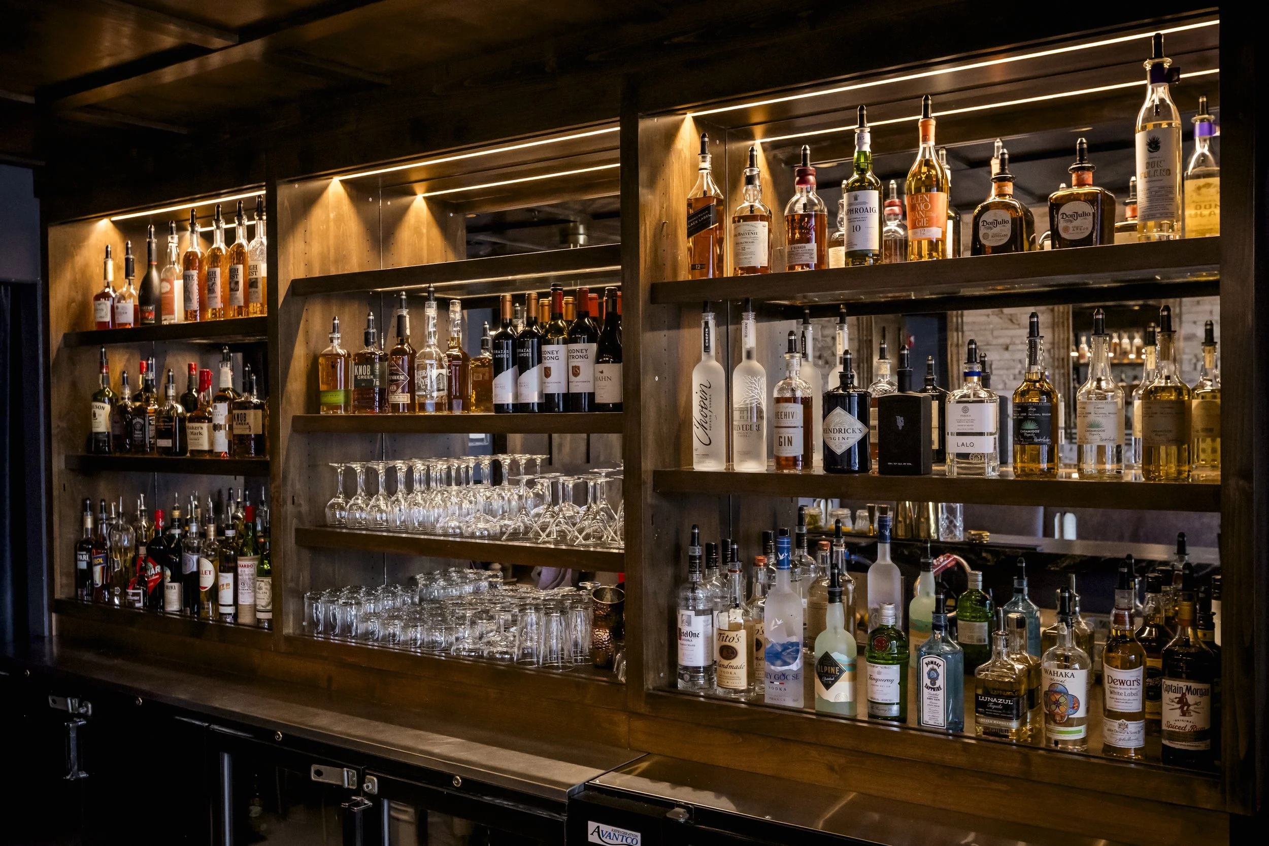 A wooden bar shelf displaying a variety of liquor bottles, with wine glasses hanging upside down below, illuminated by warm lighting.