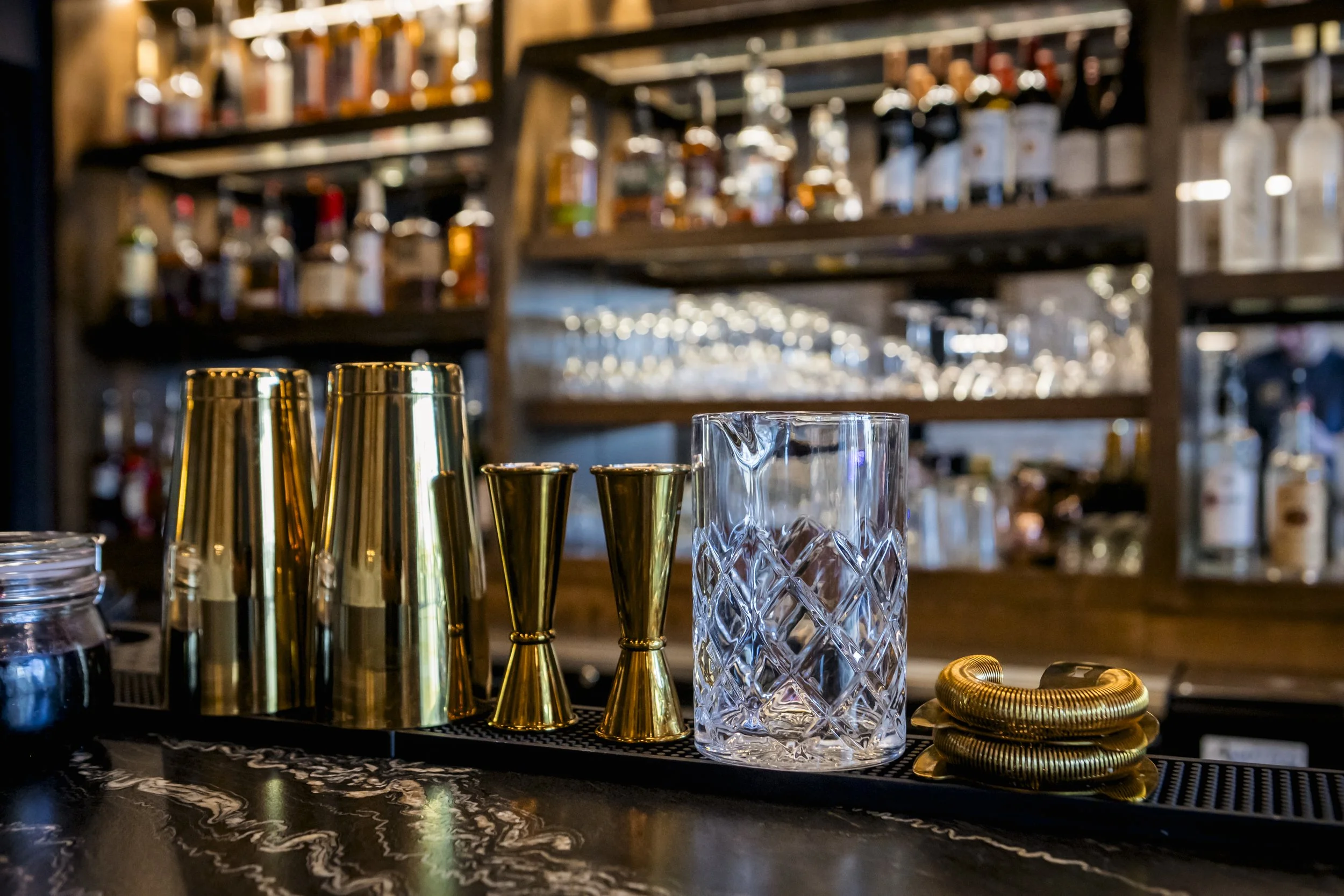 A bar counter with gold and clear cocktail shakers, a clear textured glass and gold coiled wire bracelets, with liquor bottles and glasses on shelves in the background.