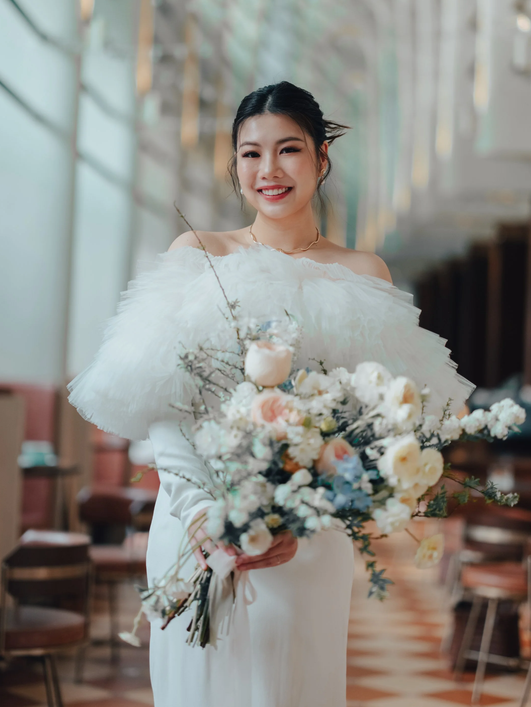 A smiling woman, Abigail Chin, an entrepreneur and content creator, in a white dress holding a large bouquet of flowers, standing inside a well-lit room with wooden chairs.