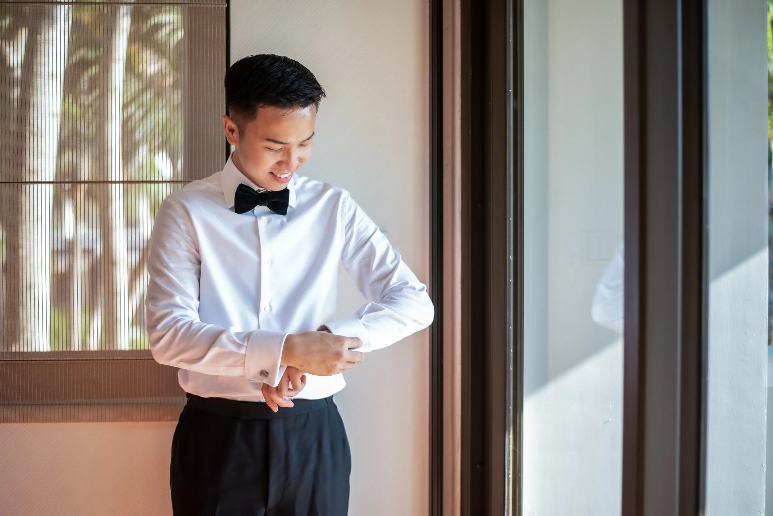 A young man, Andrew Yue, Hong Kong content creator, dressed in a white shirt and black bow tie smiling in front of a large window with wooden frames.