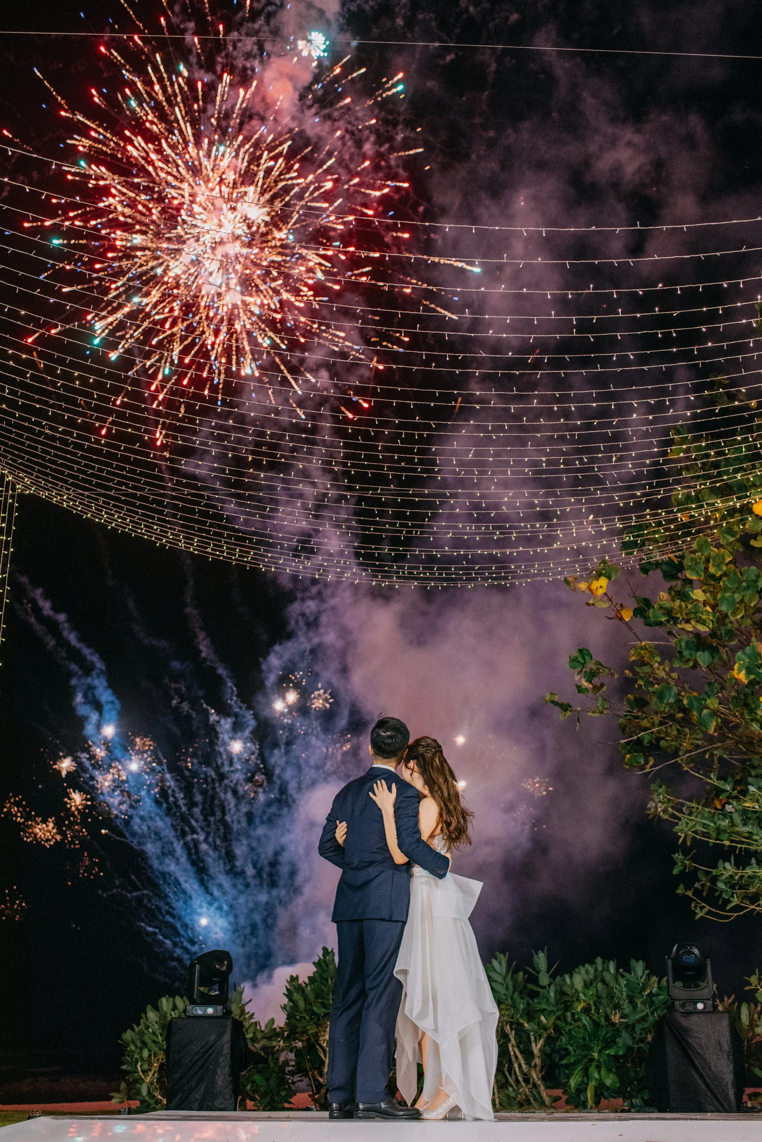 A couple, Abigail Chin & Andrew Yue, dressed in wedding attire, kissing at night under colorful fireworks and string lights.