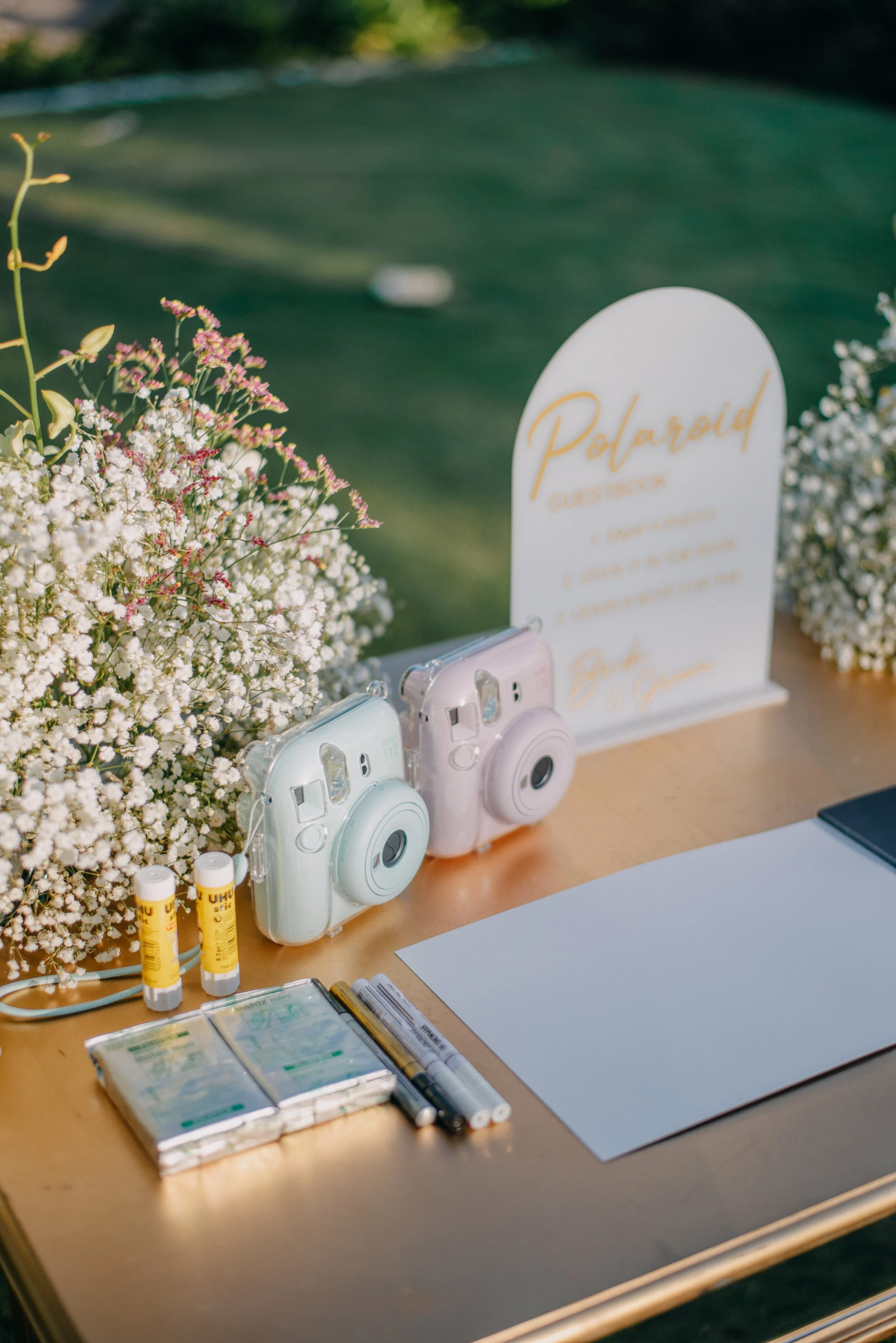 Table decorated with small cameras, white flowers, some yellow UHU glue sticks, markers, packs of tissues, a blank sheet of paper, and a sign indicating a Polaroid guestbook.
