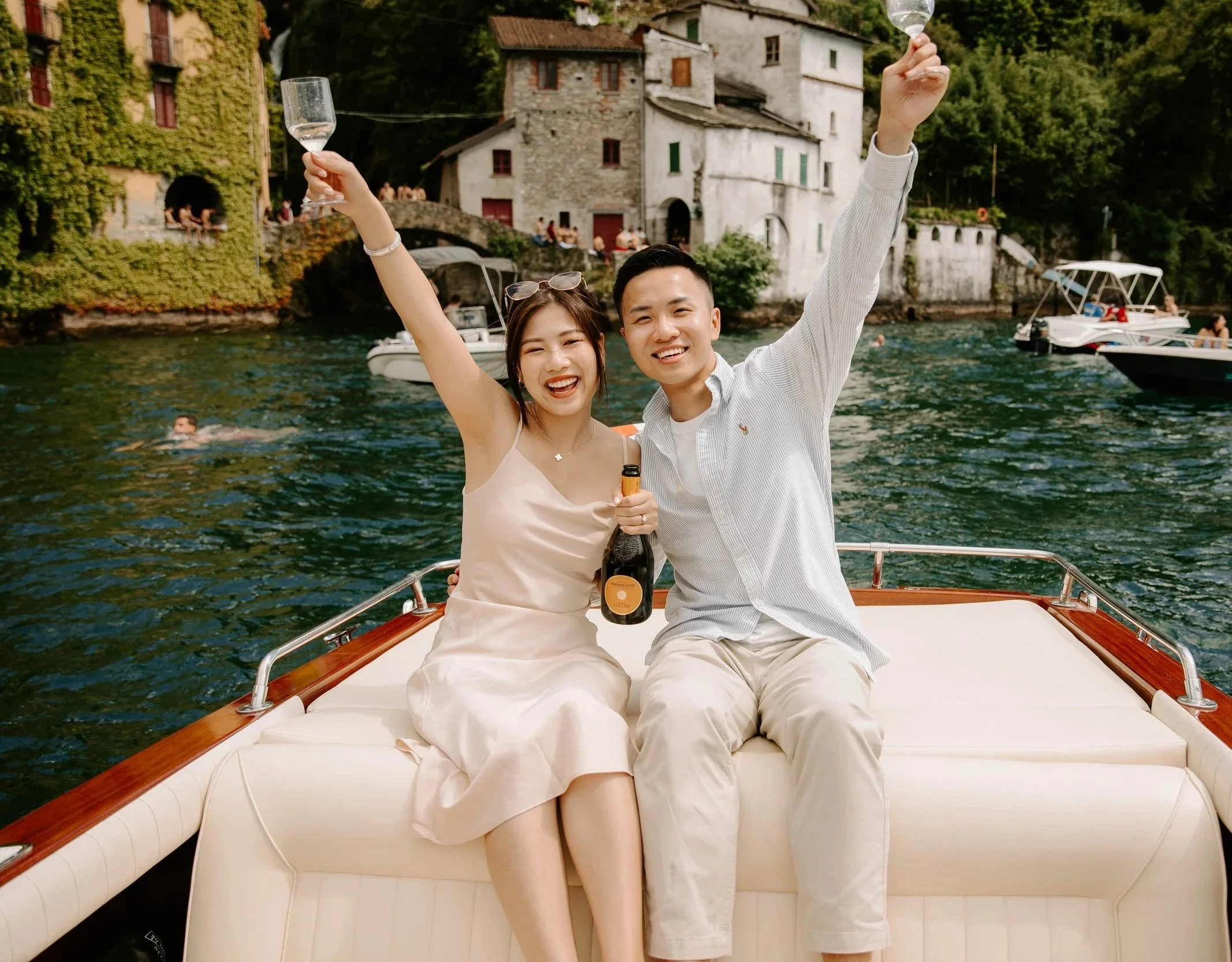 A smiling couple enjoying drinks on a boat on a river with houses and boats in the background.