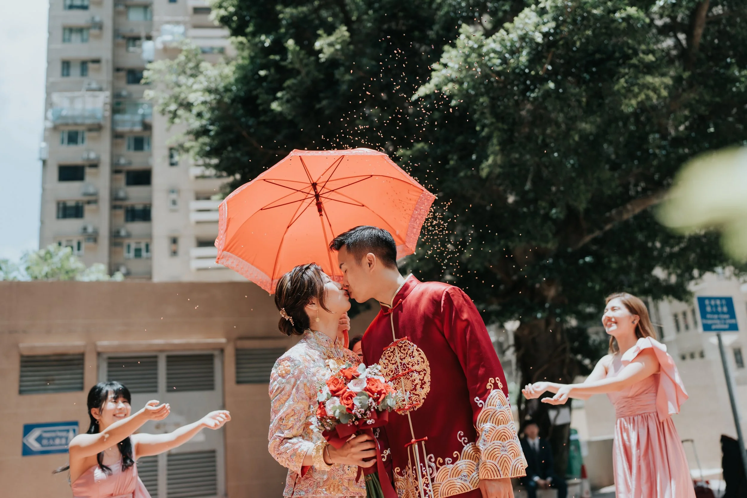 A couple dressed in traditional Chinese wedding attire sharing a kiss beneath an orange umbrella, surrounded by friends celebrating outdoors in a city setting with tall buildings and green trees.