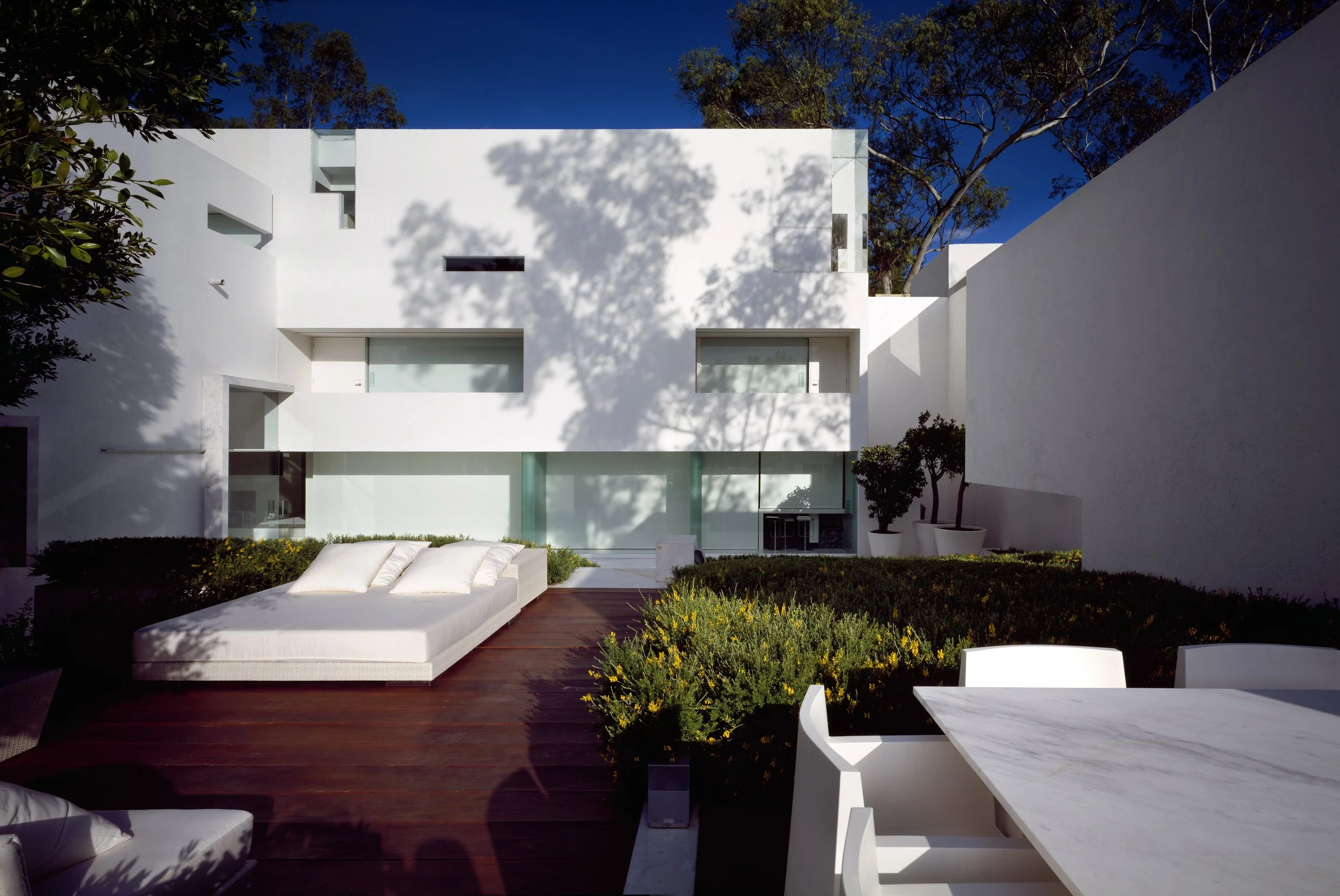 Vista de una casa moderna blanca con jardín y muebles blancos, rodeada de árboles y con cielo azul.