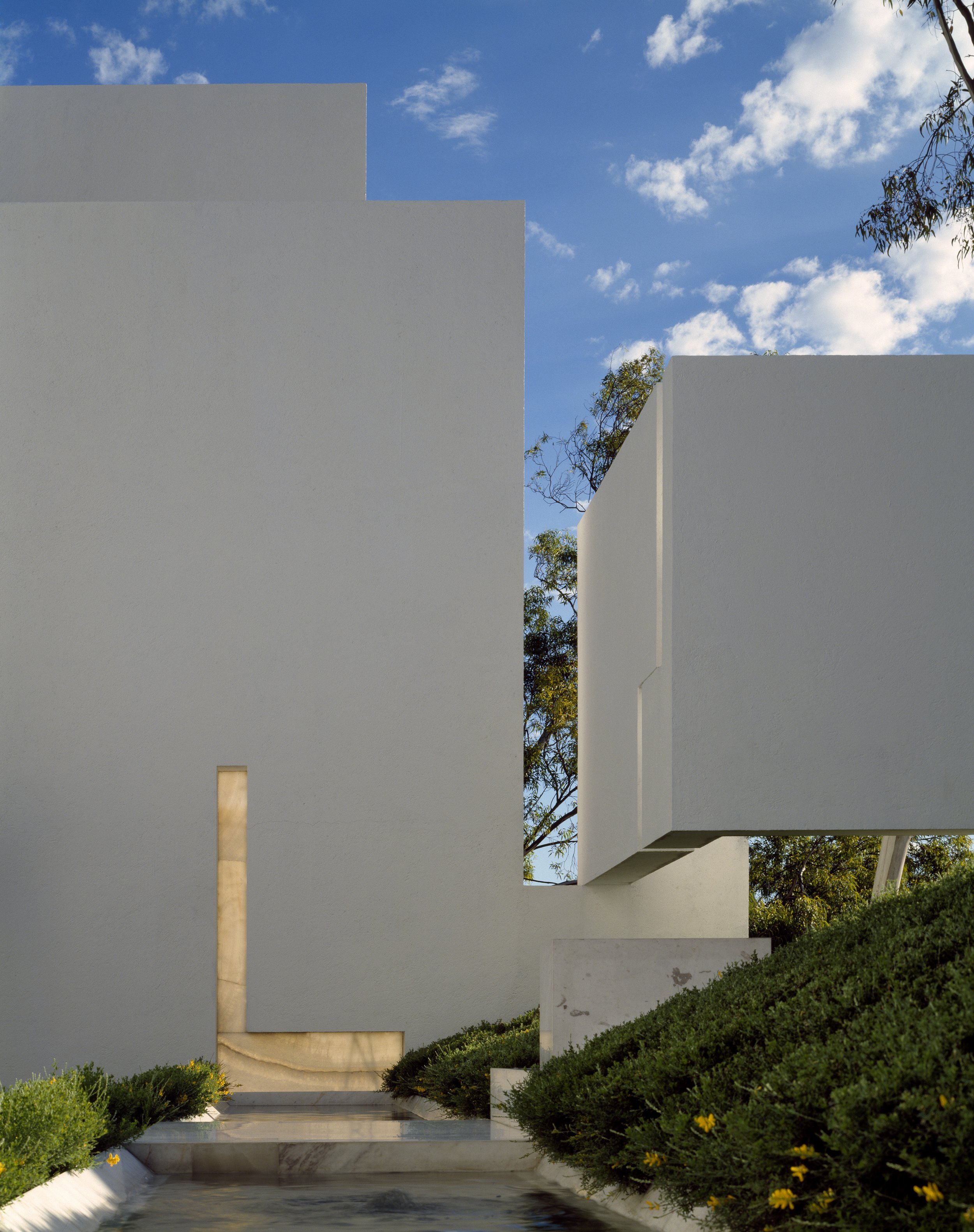 Estructuras blancas modernas con formas rectilíneas y ángulos en un espacio exterior, con vegetación y un cielo azul con nubes.