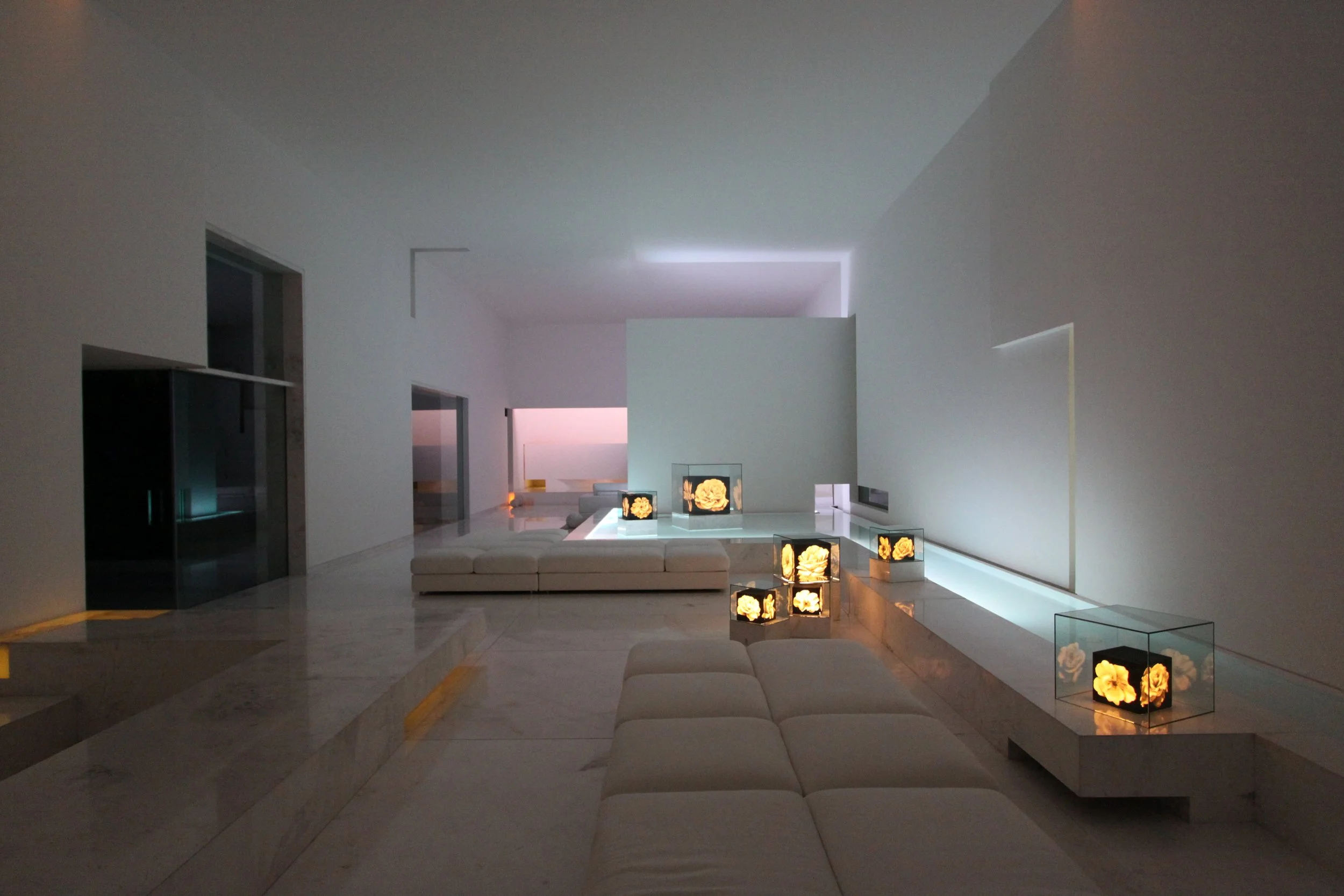 Interior de una sala moderna y minimalista con paredes blancas y muebles blancos, con pantallas que muestran flores doradas en cajas de vidrio y una área de descanso con cojines blancos.