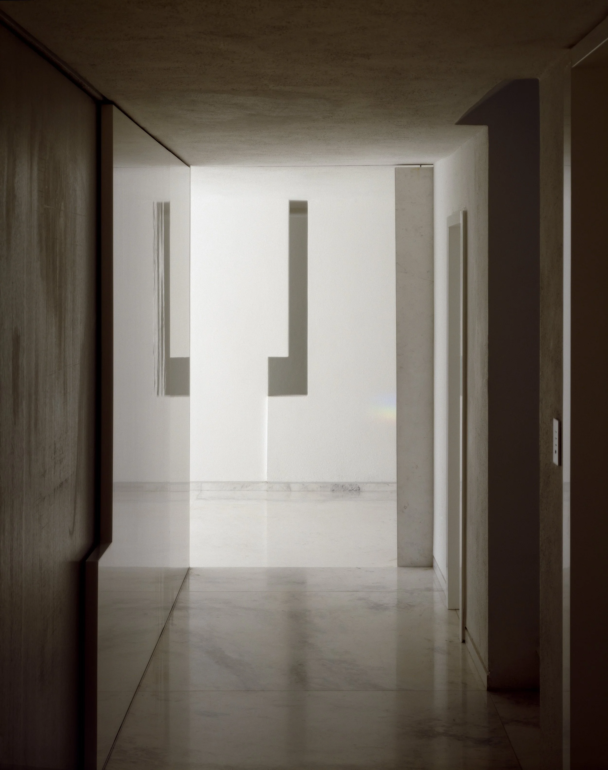 Pasillo de un edificio con paredes de color beige y piso de mármol, con dos puertas en cada extremo y un área iluminada con luz natural. Architecture & Design by Miguel Ángel Aragonés | Taller Aragonés. Photography by Joe Fletcher