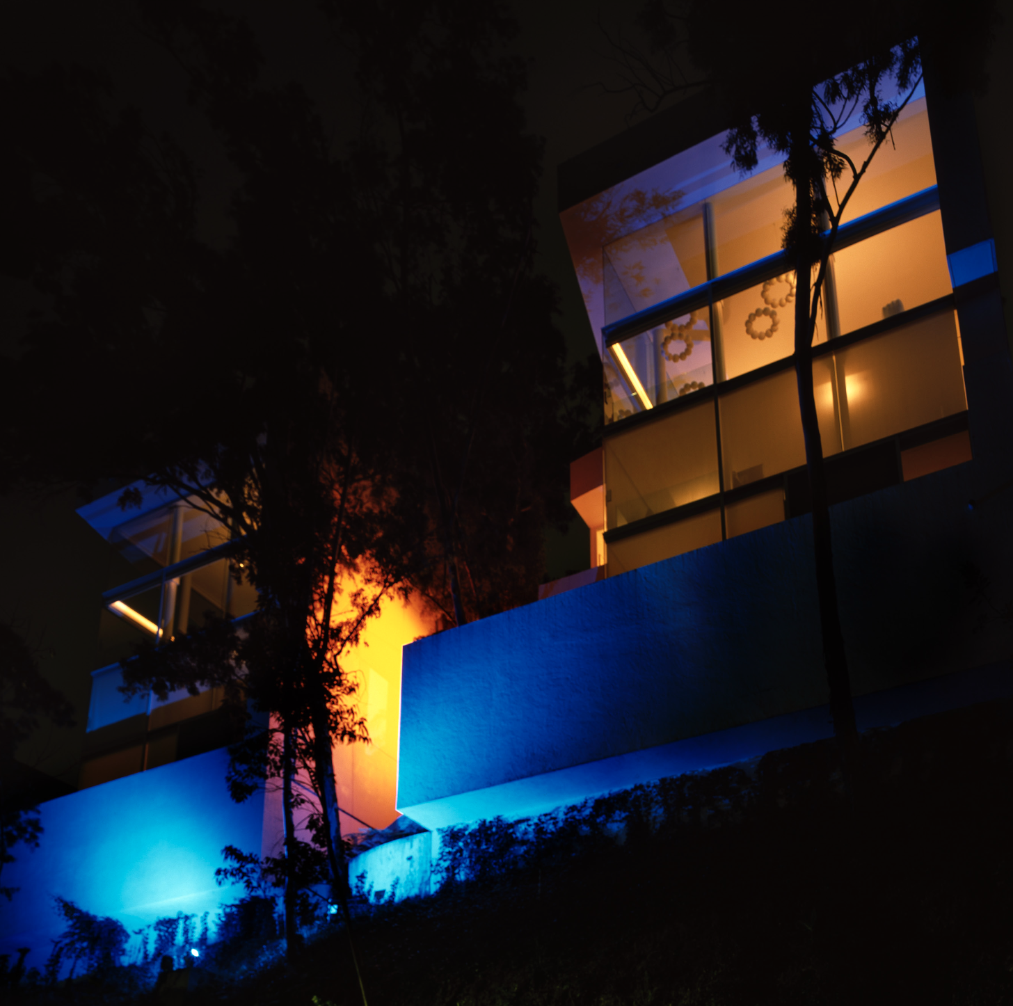 Edificio moderno con ventanas de vidrio y barandas de metal, iluminado con luces de colores azul y amarillas, rodeado de árboles en la noche.