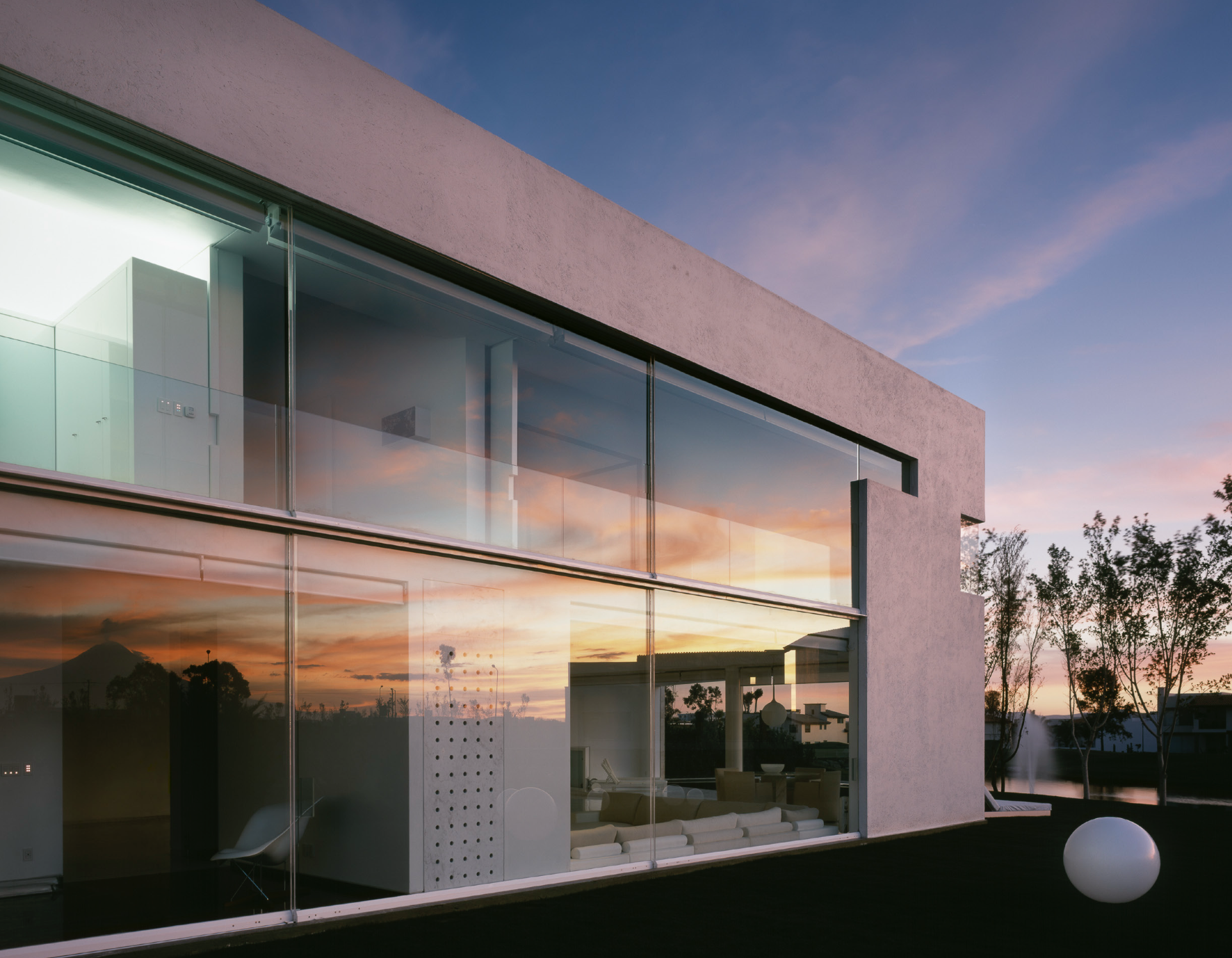 Casa moderna de dos pisos con paredes blancas y grandes ventanas de vidrio, reflejando un atardecer con cielo anaranjado y azul, rodeada de árboles y un estanque con fuentes. Casa Olivia, Architecture & Design by Miguel Ángel Aragonés | Taller Aragon