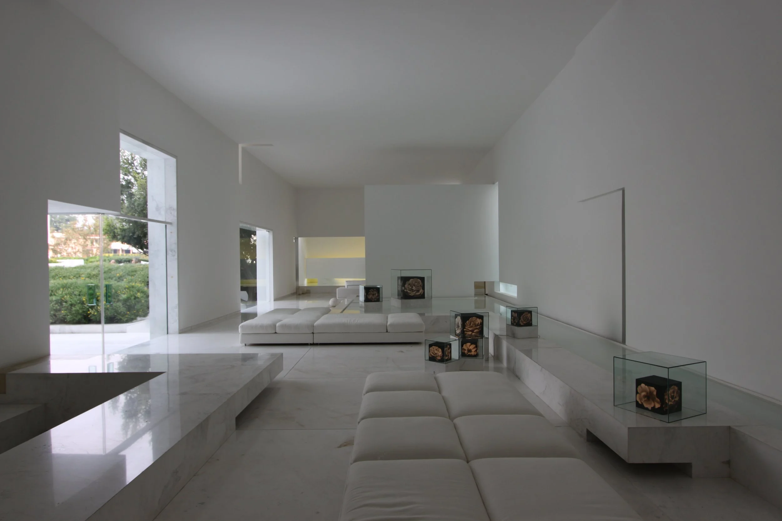 Interior moderna y minimalista con paredes y techo blancos, grandes ventanas, sillones blancos y mesas con decoraciones florales en cajas de acrílico