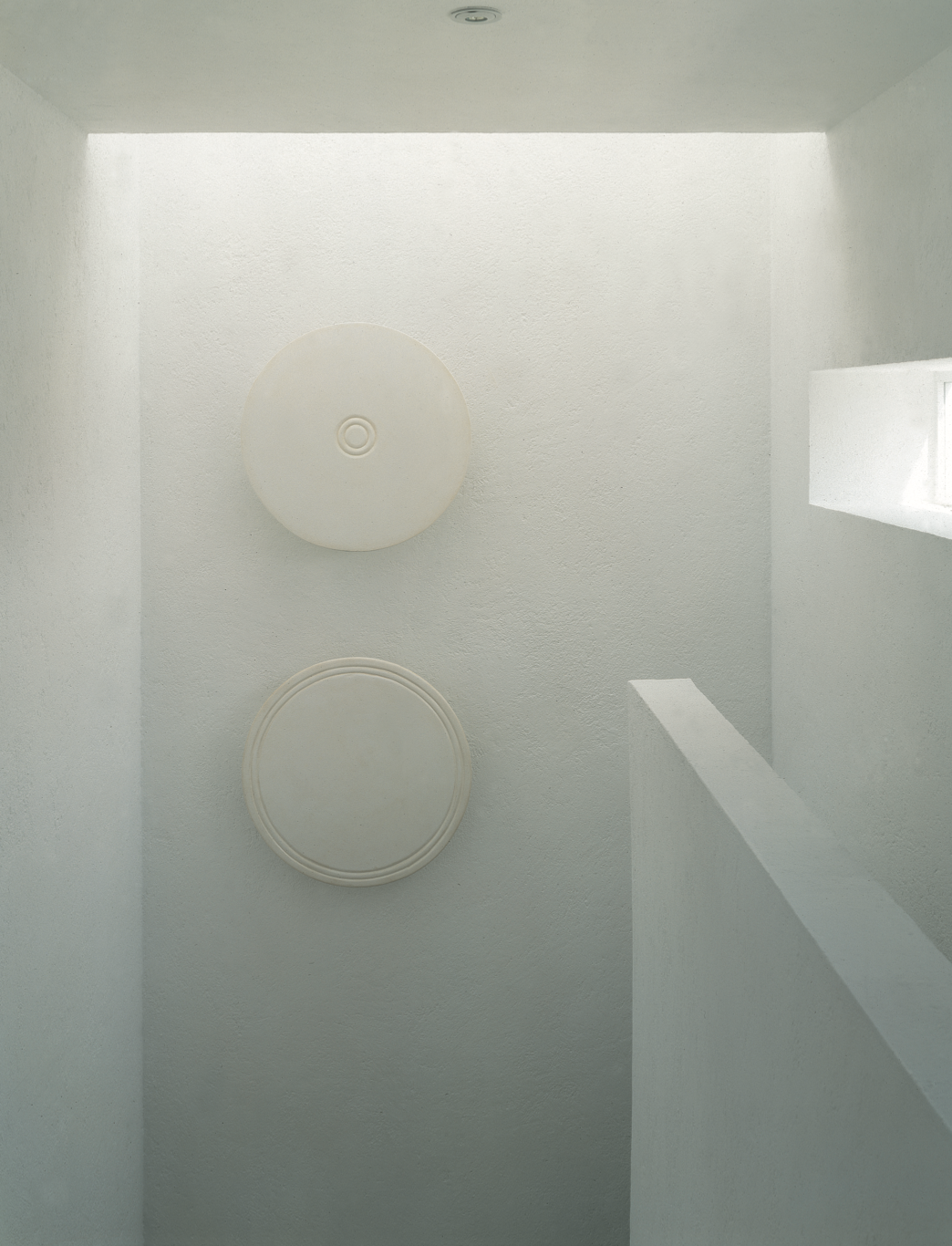 Imagen minimalista de un techo blanco con dos bocinas circulares empotradas, una encima de la otra, y una pequeña ventana rectangular por donde entra luz natural.