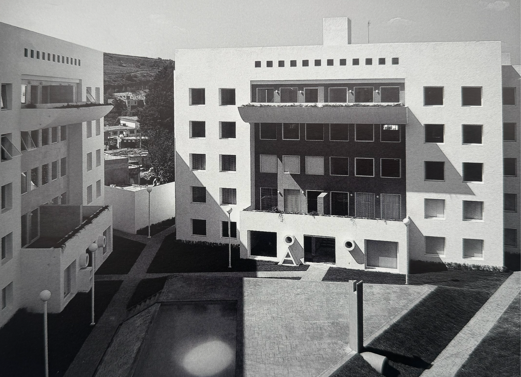 Vista en blanco y negro de edificios modernos con ventanas cuadradas, una terraza con barandilla y áreas de sombra, y farolas en el patio central.